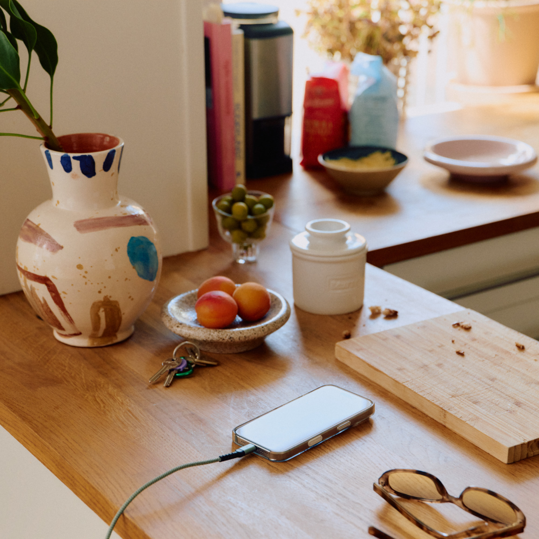A busy wooden kitchen counter with a vase, apricots, olives, keys, a charging phone, and sunglasses.