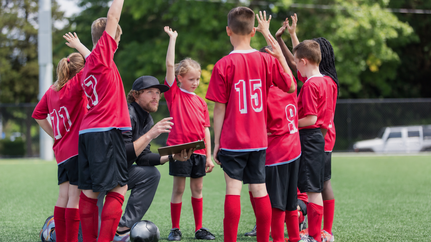 Young soccer players in red jerseys huddle around their coach, some with hands raised, on a green field.