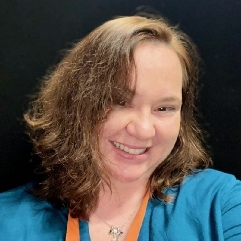A smiling woman with brown, wavy hair wears a blue top, an orange lanyard, and a cross necklace against a dark background.