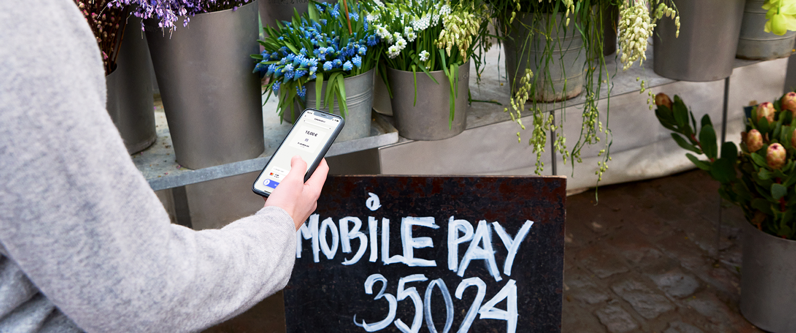 A person uses a smartphone to make a mobile payment at a flower stall with a "Mobile Pay" sign.