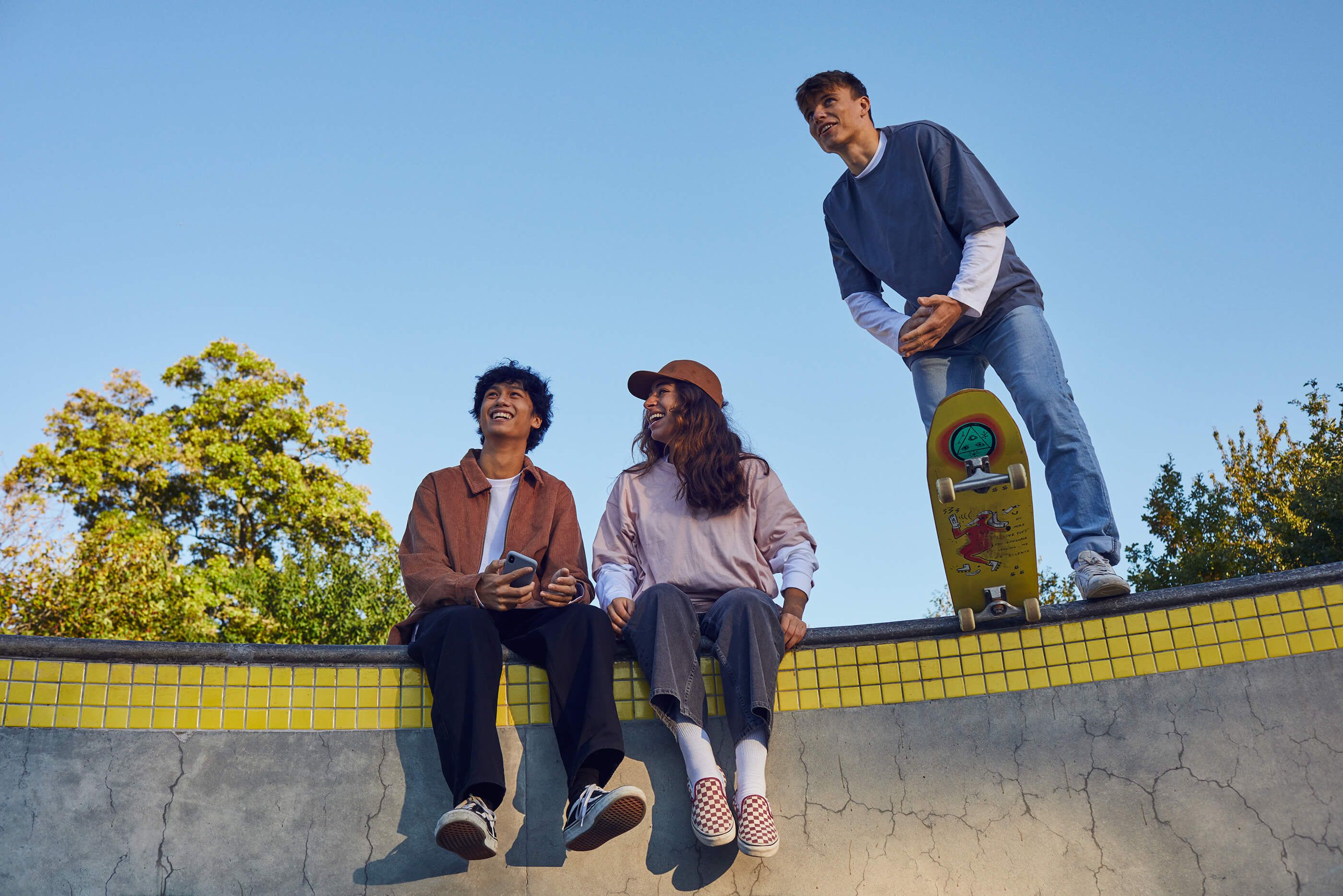 Three smiling friends at a skate park: two sit on a yellow-tiled wall, one stands with a skateboard.