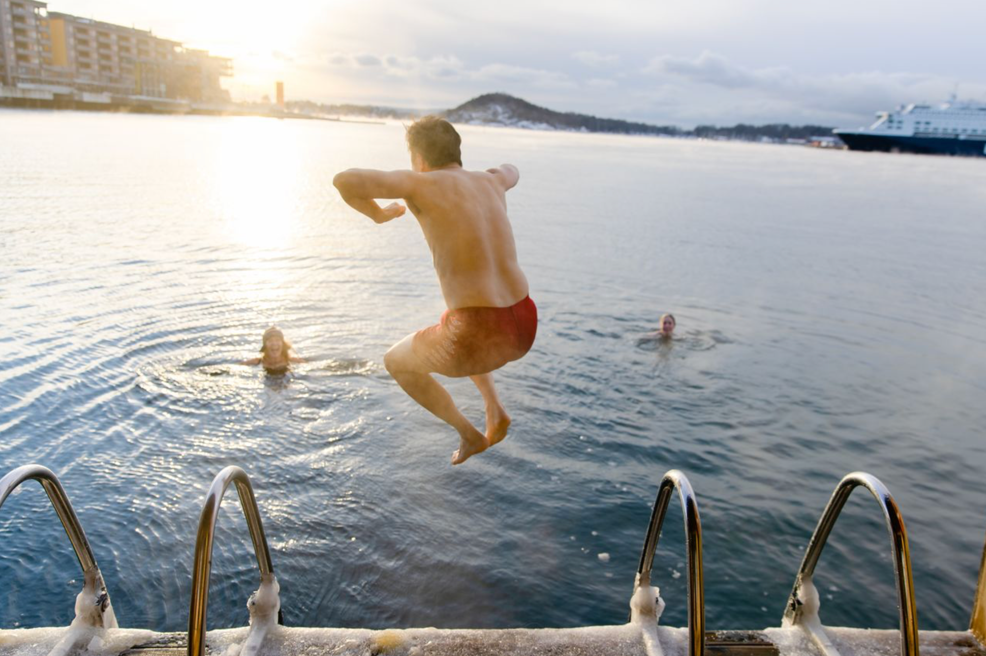 A man in red swim trunks jumps into cold water from a dock with icy steps, as two other people swim nearby under a bright sky.