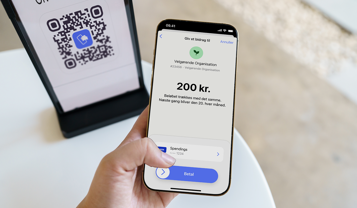 A hand holds a smartphone displaying a 200 kr. donation payment to a charitable organization, with a QR code stand in the background.