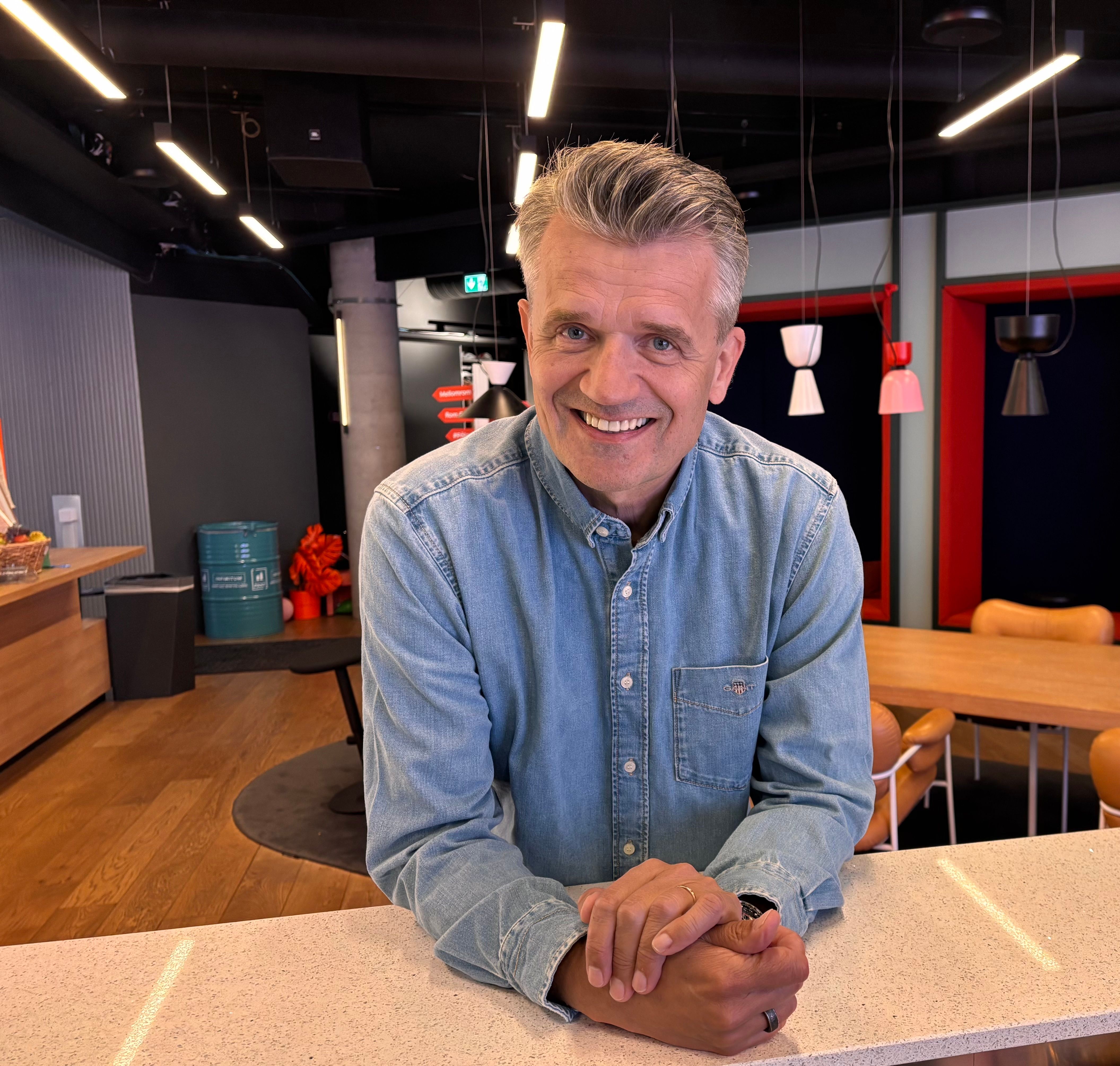 Smiling man with grey hair in a denim shirt leans on a counter in a modern interior.