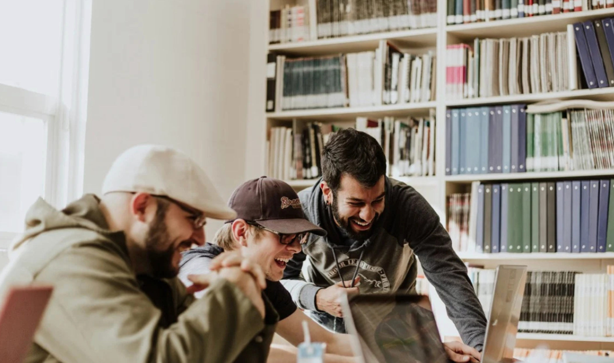 Three men laughing over a laptop in a library.