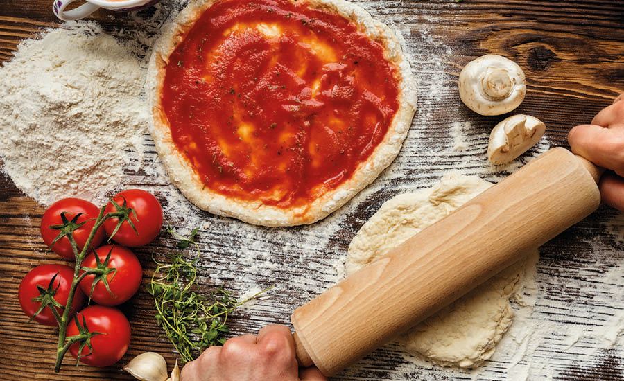 Hands rolling pizza dough on a floured wooden table with a sauced pizza base, tomatoes, mushrooms, and herbs.