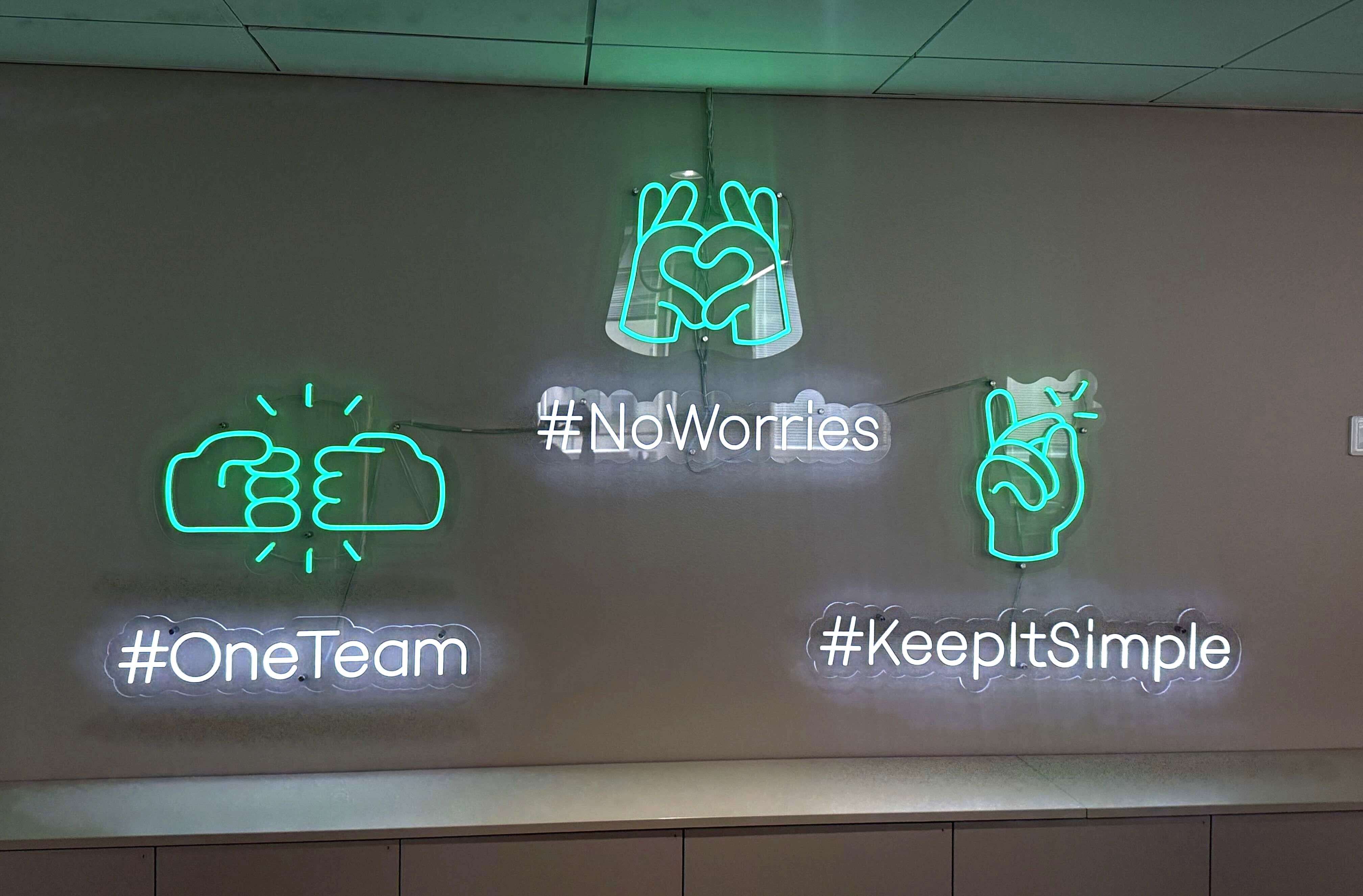 Neon signs on a wall with teal icons and white text: a heart made of hands above "#NoWorries", a fist bump above "#OneTeam", and a peace sign above "#KeepItSimple".