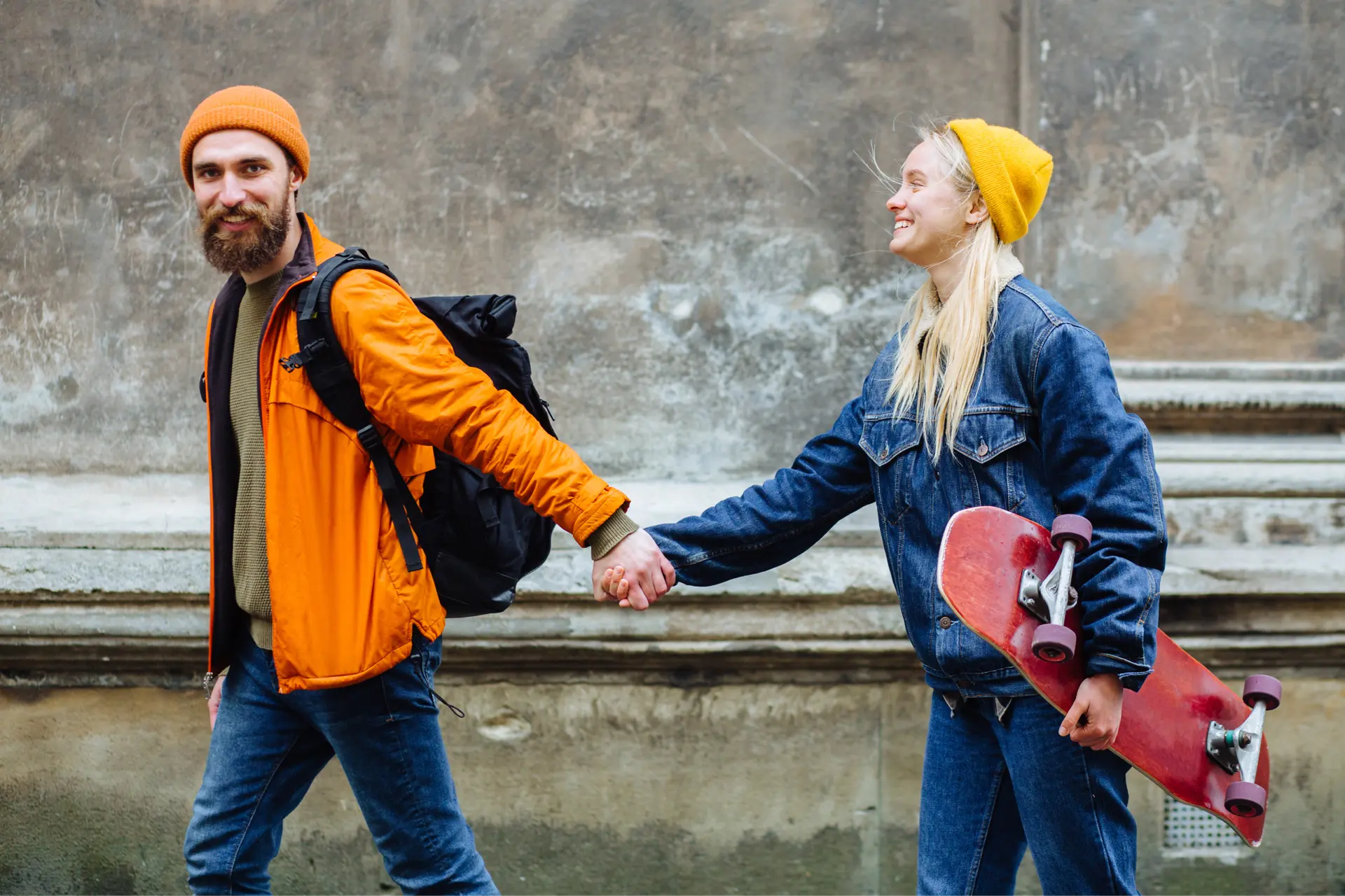 A happy couple walks hand-in-hand, the man wearing an orange jacket and the woman in a denim jacket carrying a red skateboard.