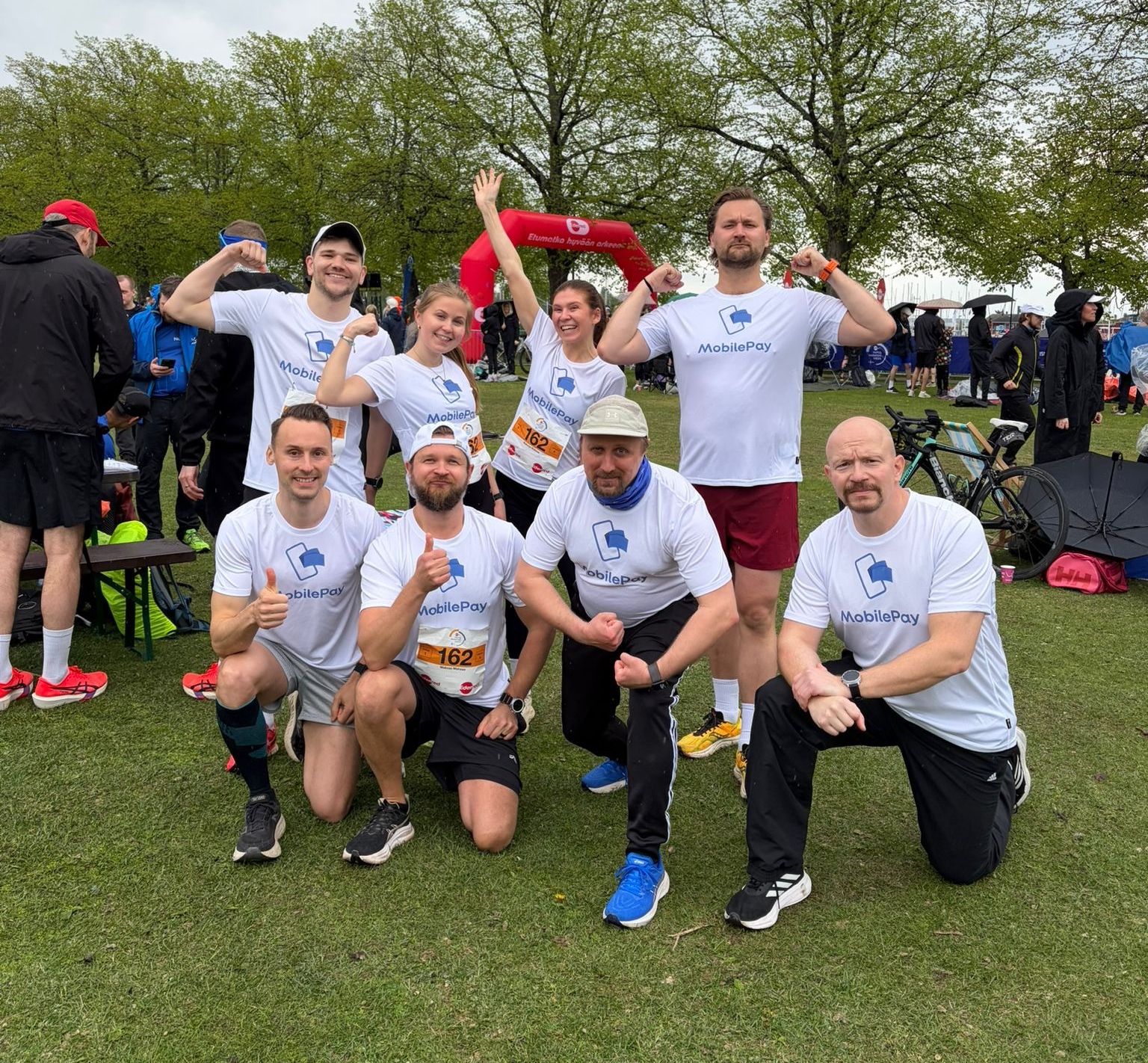 Eight people in white MobilePay t-shirts pose on a grassy field, some flexing muscles, with a red arch in the background.