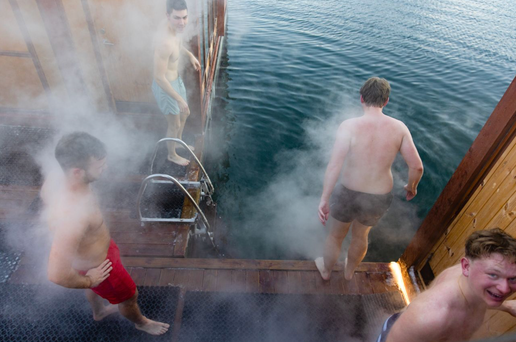 Four men on a wooden pier, enveloped in steam, as they enter or prepare to enter cold water.