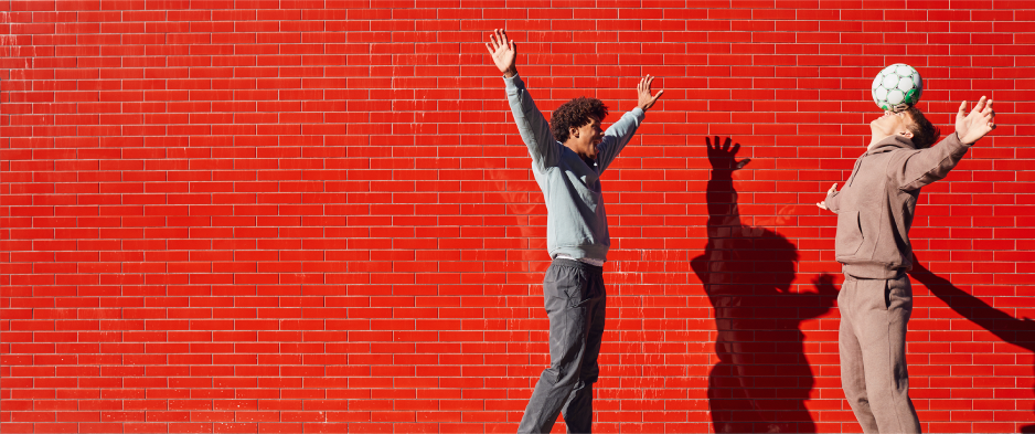 Two people against a red brick wall; one balances a soccer ball on their head, the other raises their arms.