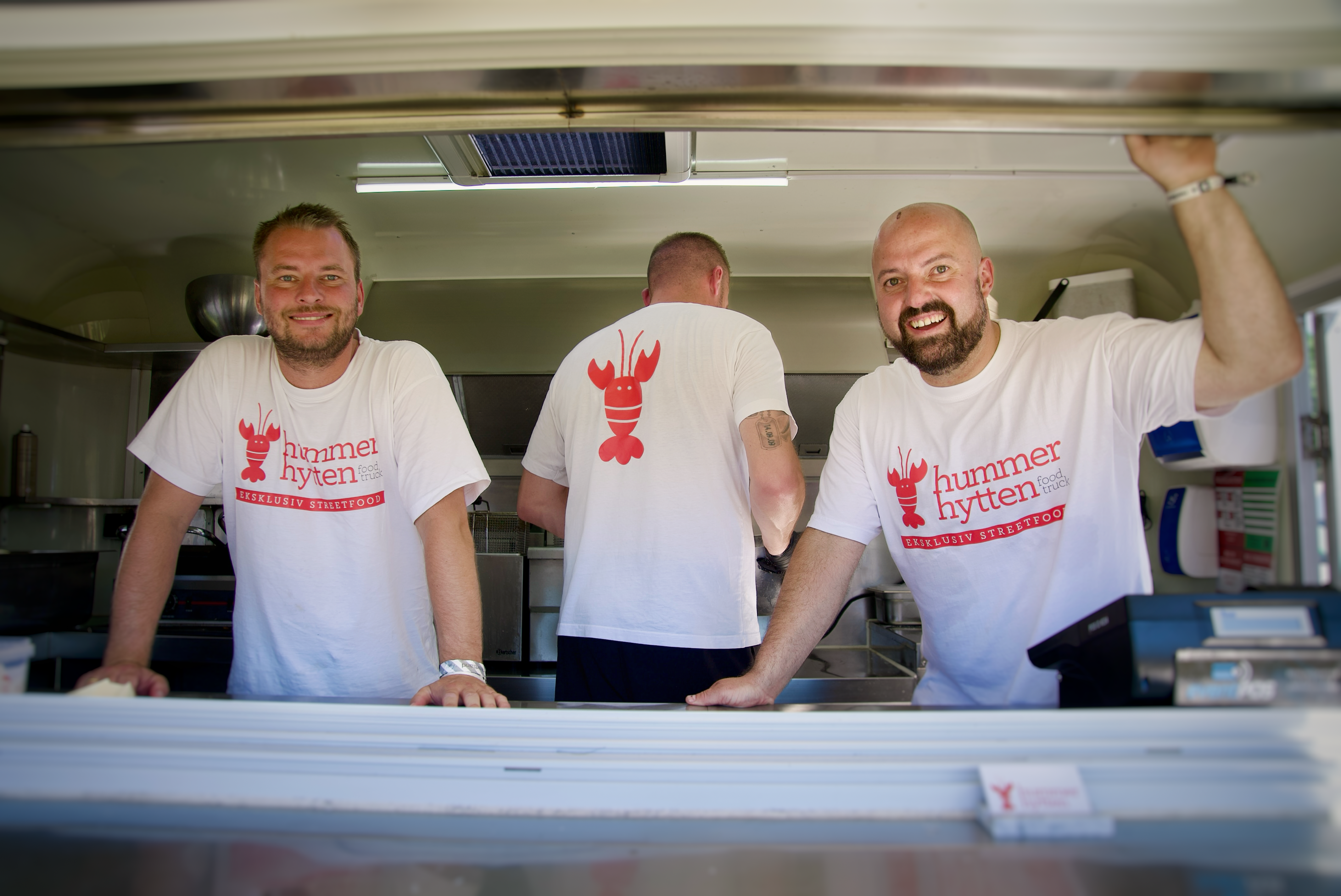 Two smiling men in white t-shirts with a red lobster logo and "Hummer Hytten" text stand in a food truck, while a third man faces away.