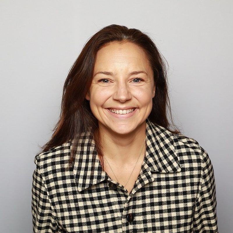 A smiling woman with long brown hair wearing a black and white checkered shirt.