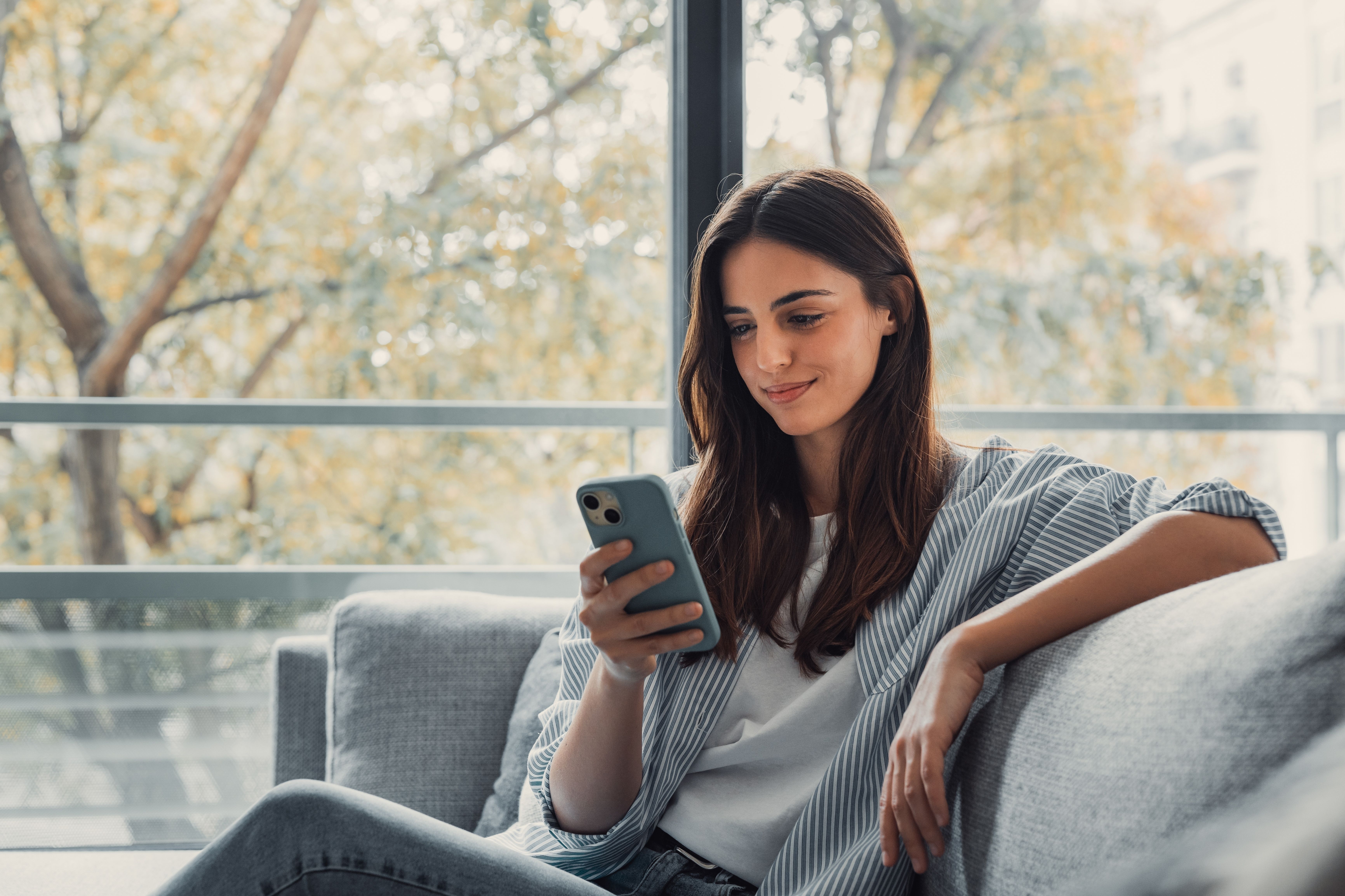 A woman on a couch smiles while looking at her smartphone.