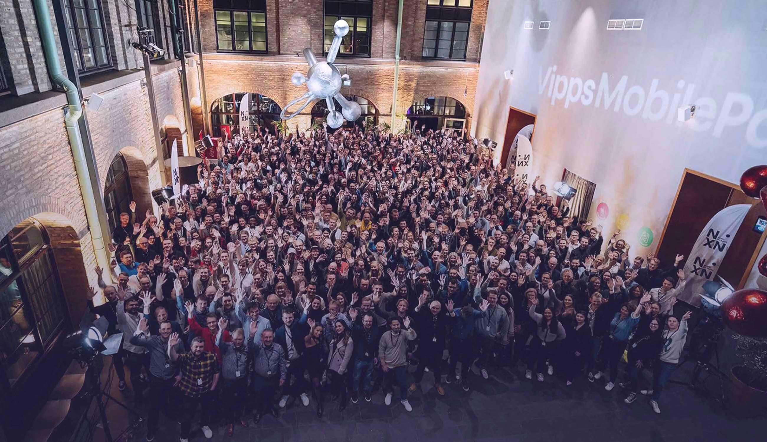 A large group of people waving in an outdoor courtyard, with "Vipps MobilePo" projected on a building and an abstract sculpture hanging overhead.