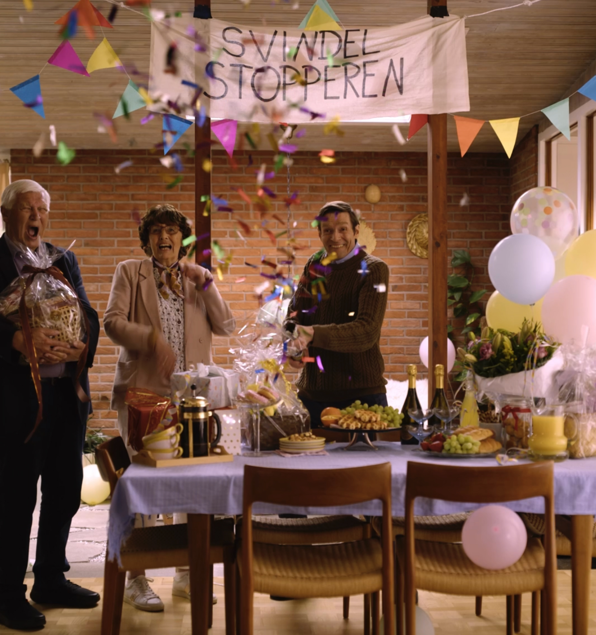 An older man, an older woman, and a younger man joyfully celebrate with falling confetti and a banner reading "FRAUD STOPPER" above a table laden with gifts and food.