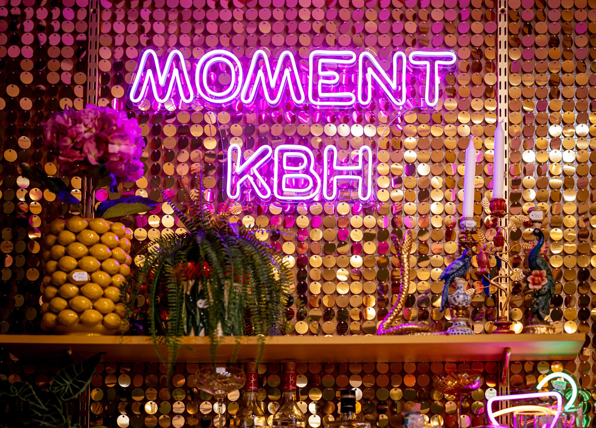 Pink neon sign "MOMENT KBH" on a gold sequin wall, above a shelf with lemons, plants, and decorative candles.
