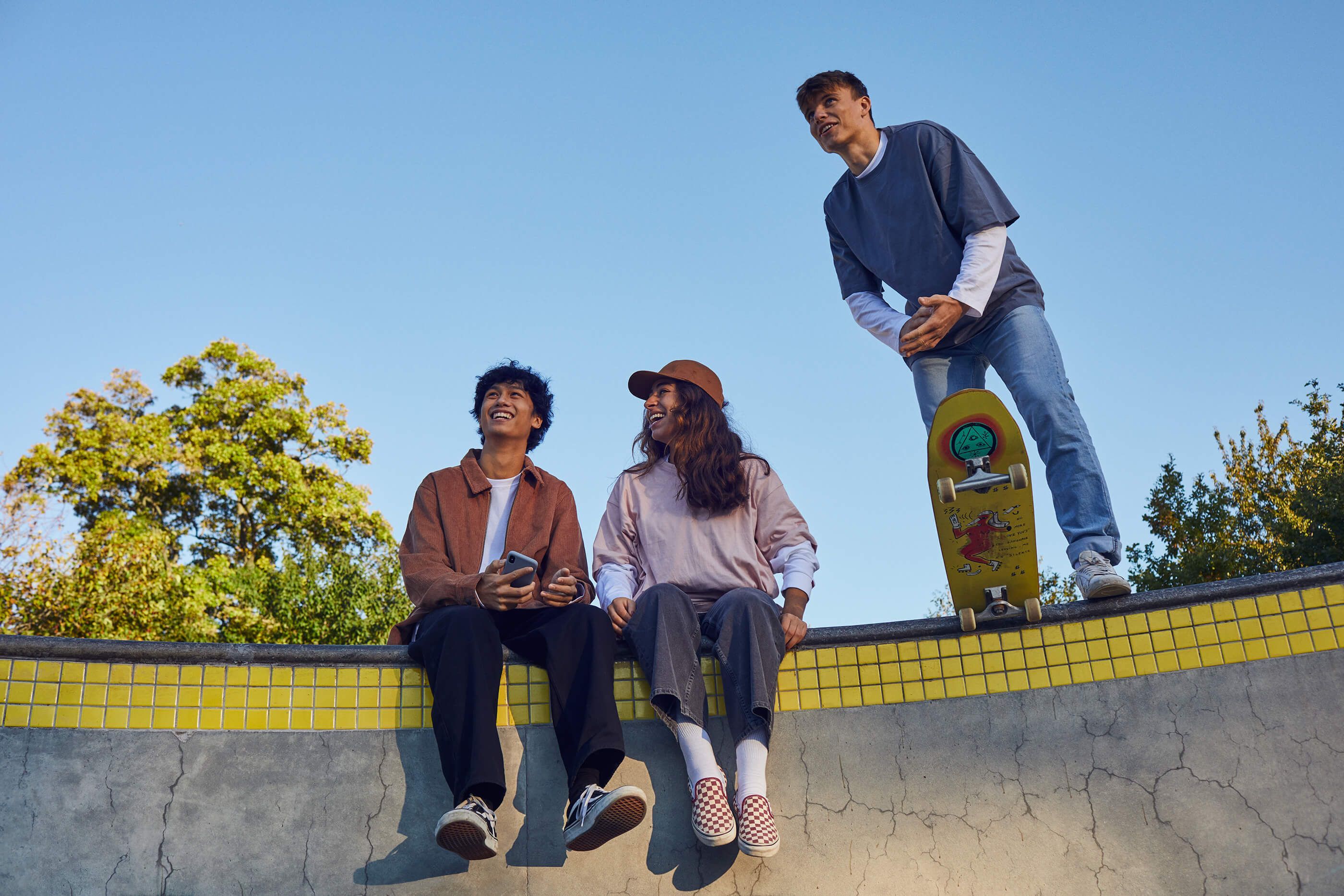 Three friends smiling in a skatepark, two sitting on the edge and one standing on a skateboard.