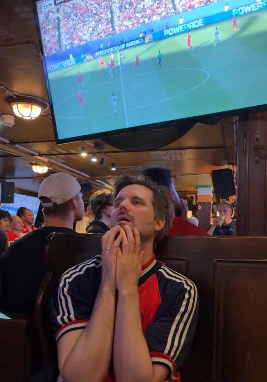 A man with hands clasped under his chin intently watches a soccer game on a large TV in a dimly lit pub.