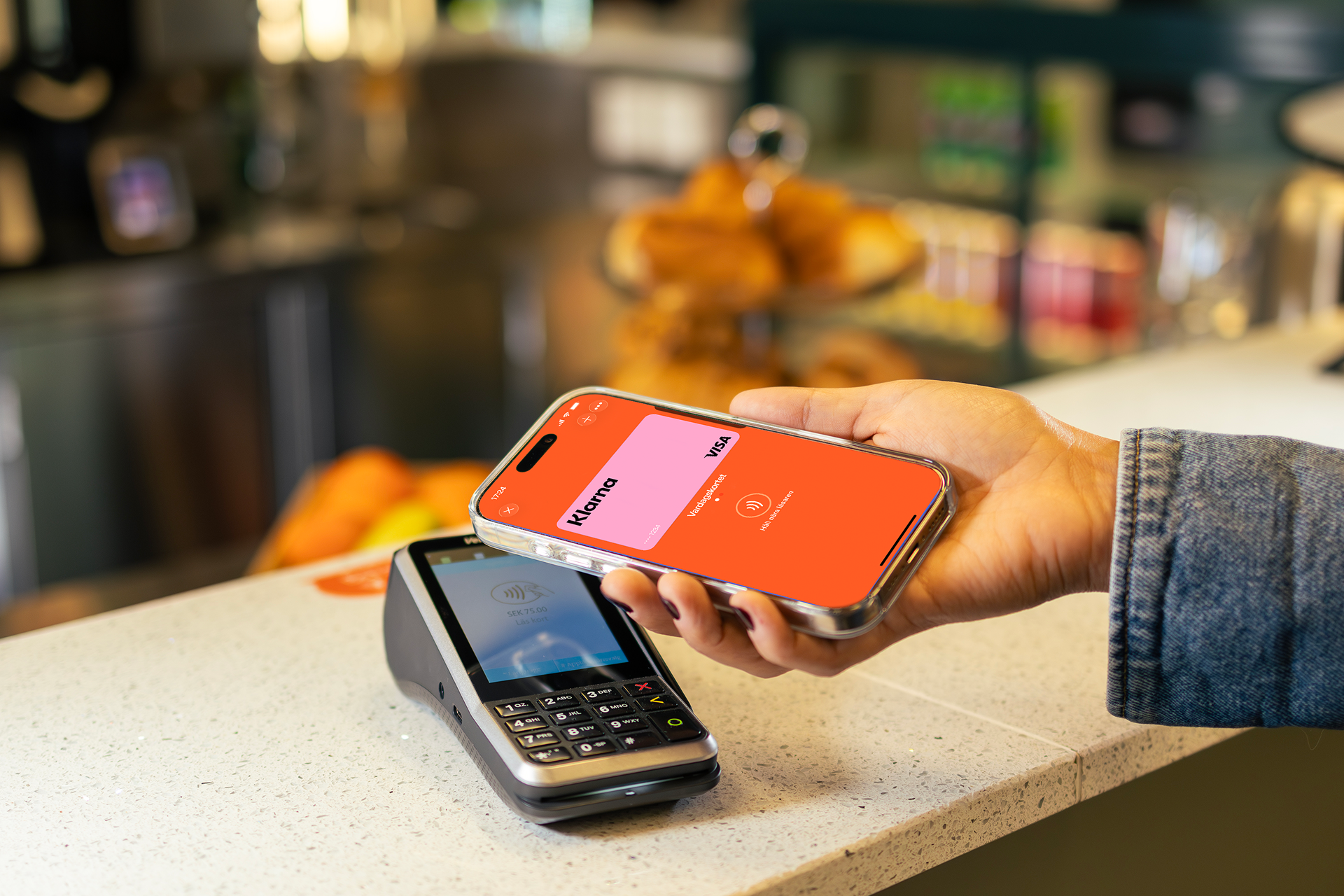 A hand holds a smartphone displaying the Vipps app with a Klarna card over a payment terminal for a contactless transaction.