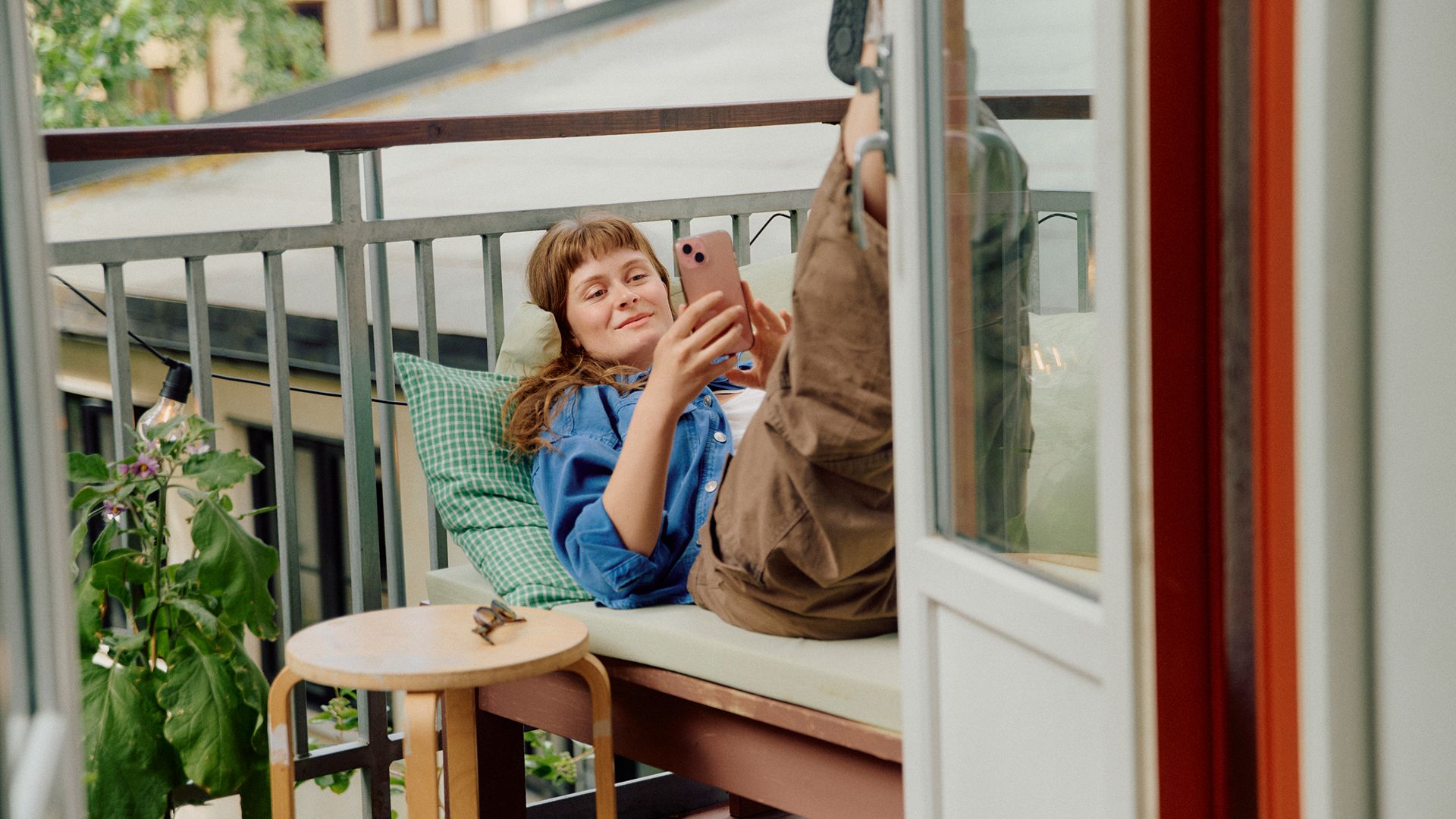 A woman lies relaxed on a balcony bench, smiling at her phone with her legs propped up.