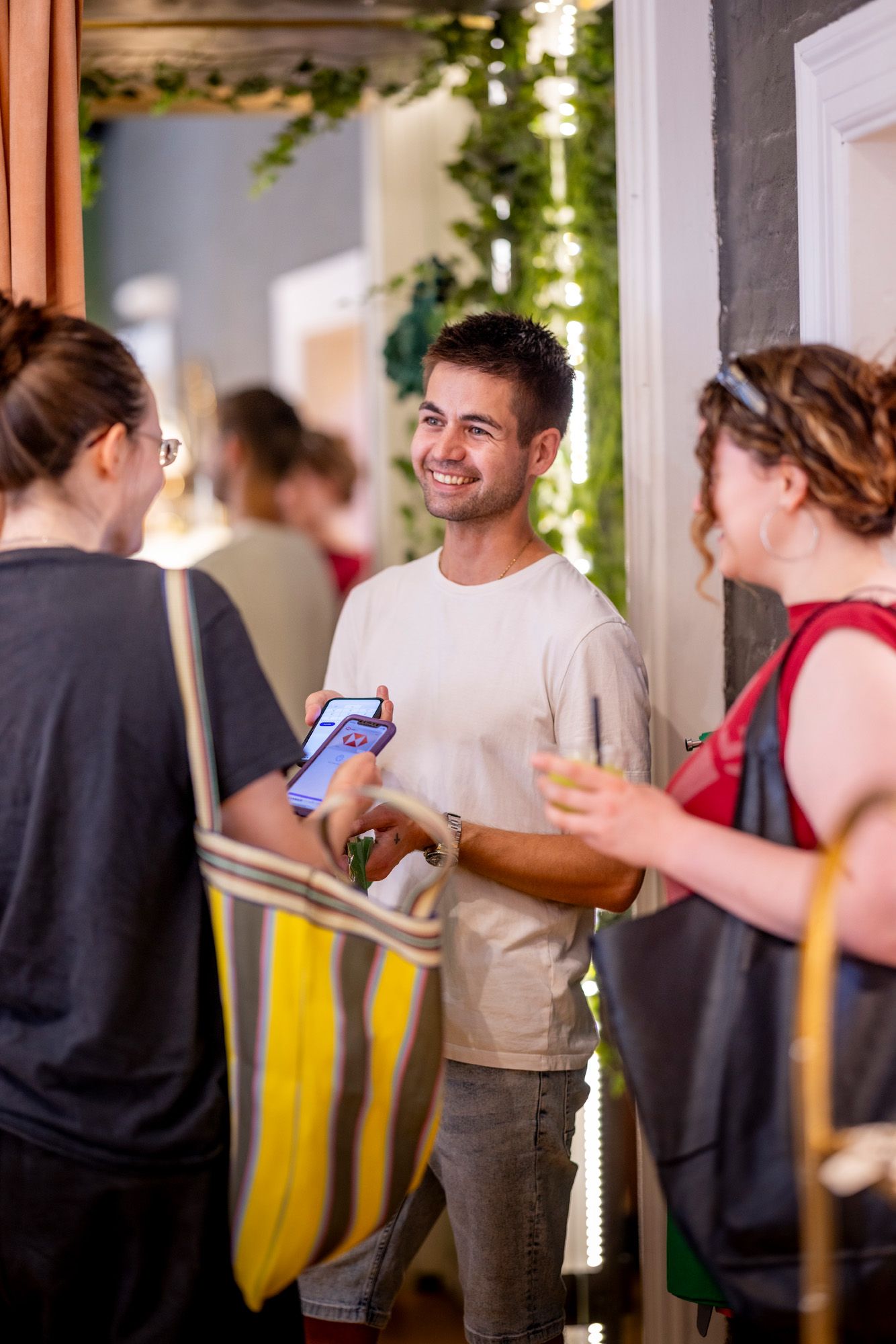 A smiling man holds two smartphones, showing them to two women in an indoor setting. One woman carries a striped tote bag, the other holds a drink.
