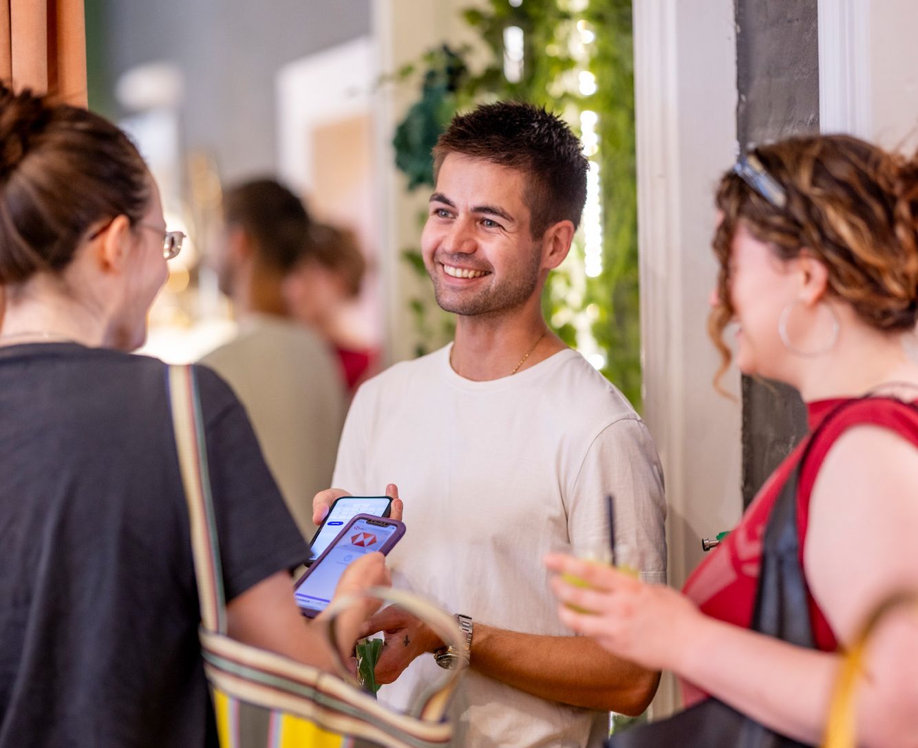 A smiling man holds two phones while talking to two women, one holding a drink, at an indoor event.