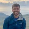A man is smiling at the camera with a blue zipped up hoody. There is nature behind him.