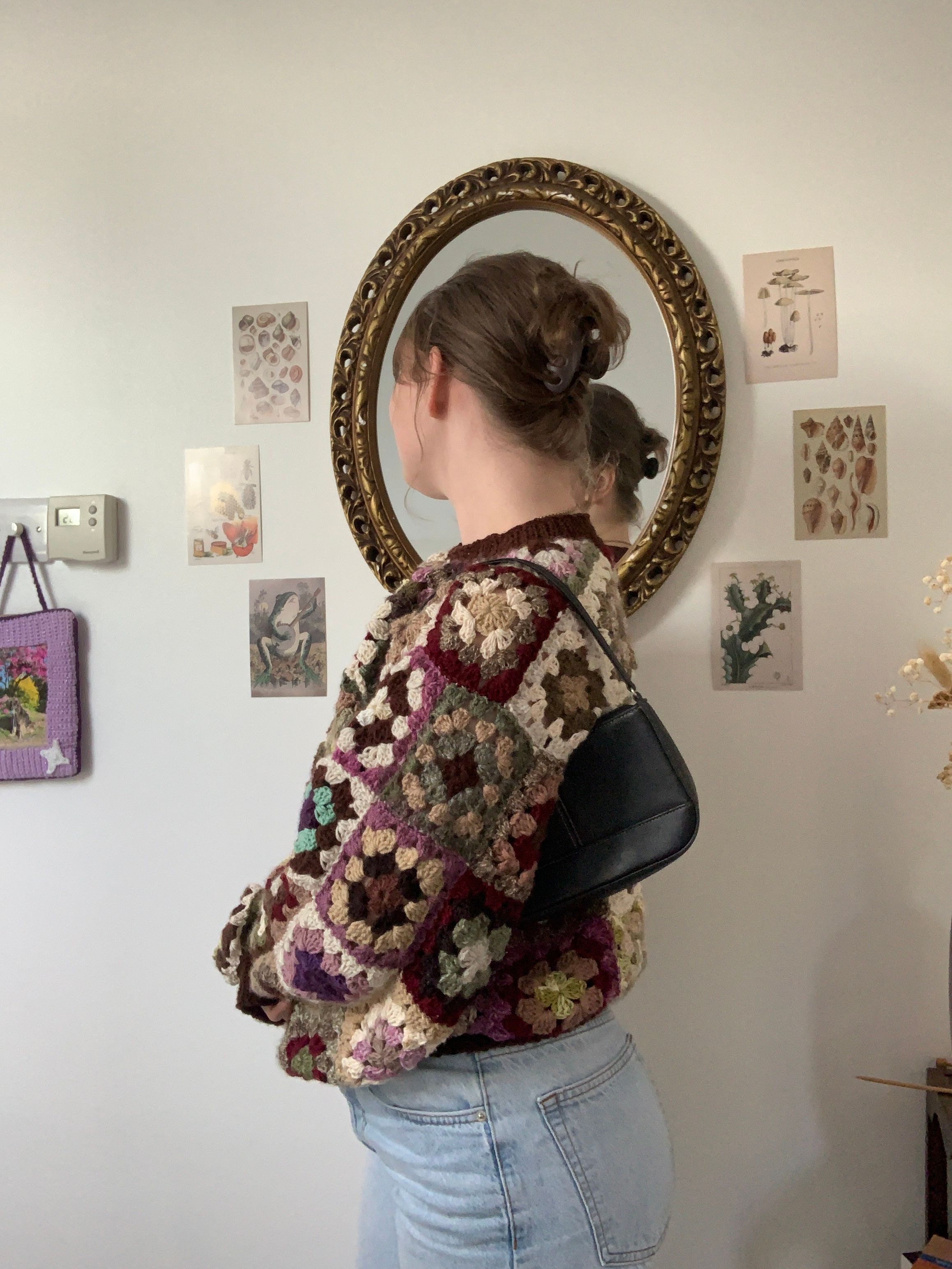 Granny square sweater worn with jeans and a mini shoulder bag. A vintage mirror sits in the background with cottagecore style wall prints.