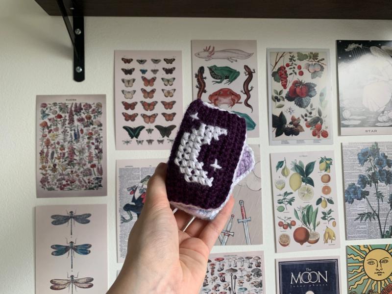 Finished purple spell book with a white cross-stitched crescent moon, held up in front of a wall of prints