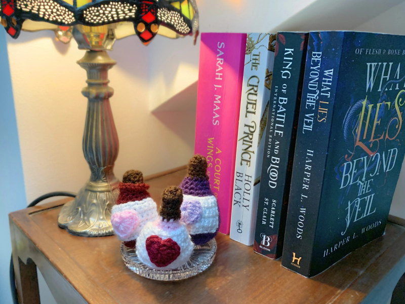 3 mini love potions sitting on trinket tray, which sits on a bedside table. A tiffany lamp lights up the scene and a stack of fantasy books sits on the wooden table.