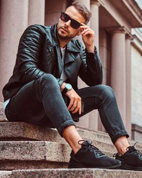 A man in a leather jacket and sunglasses witting on stone steps in front of stone columns