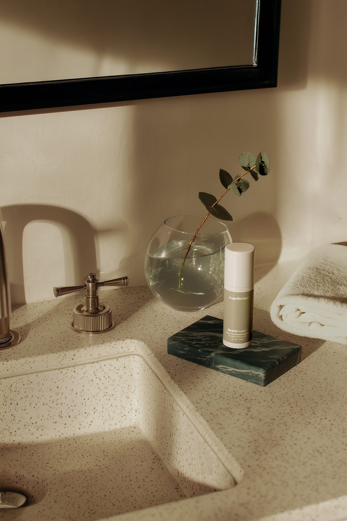 KapelleSkin moisturizer beside glass and a budding stem at a bathroom sink.