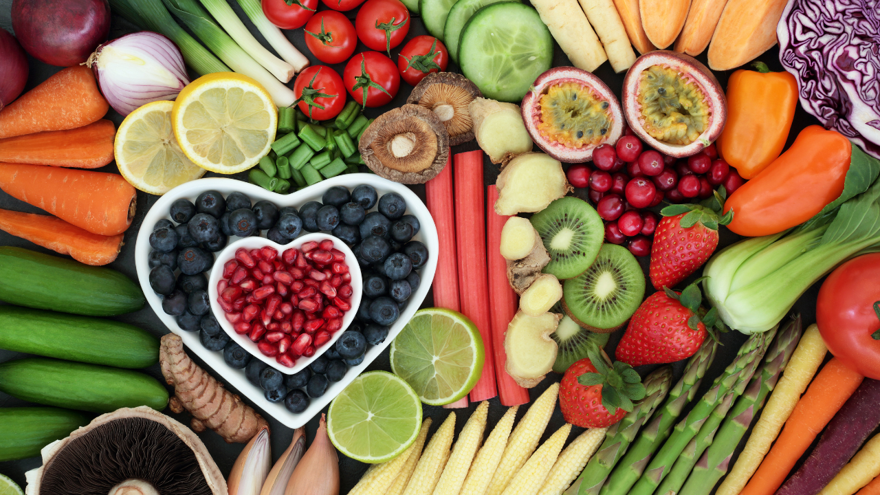 Overhead arrangement of colorful fresh fruits and vegetables surrounding a heart-shaped bowl filled with blueberries and pomegranate seeds.