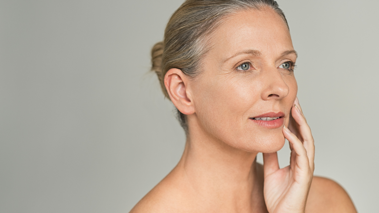 Mature woman with gray hair touching her cheek against a neutral background.