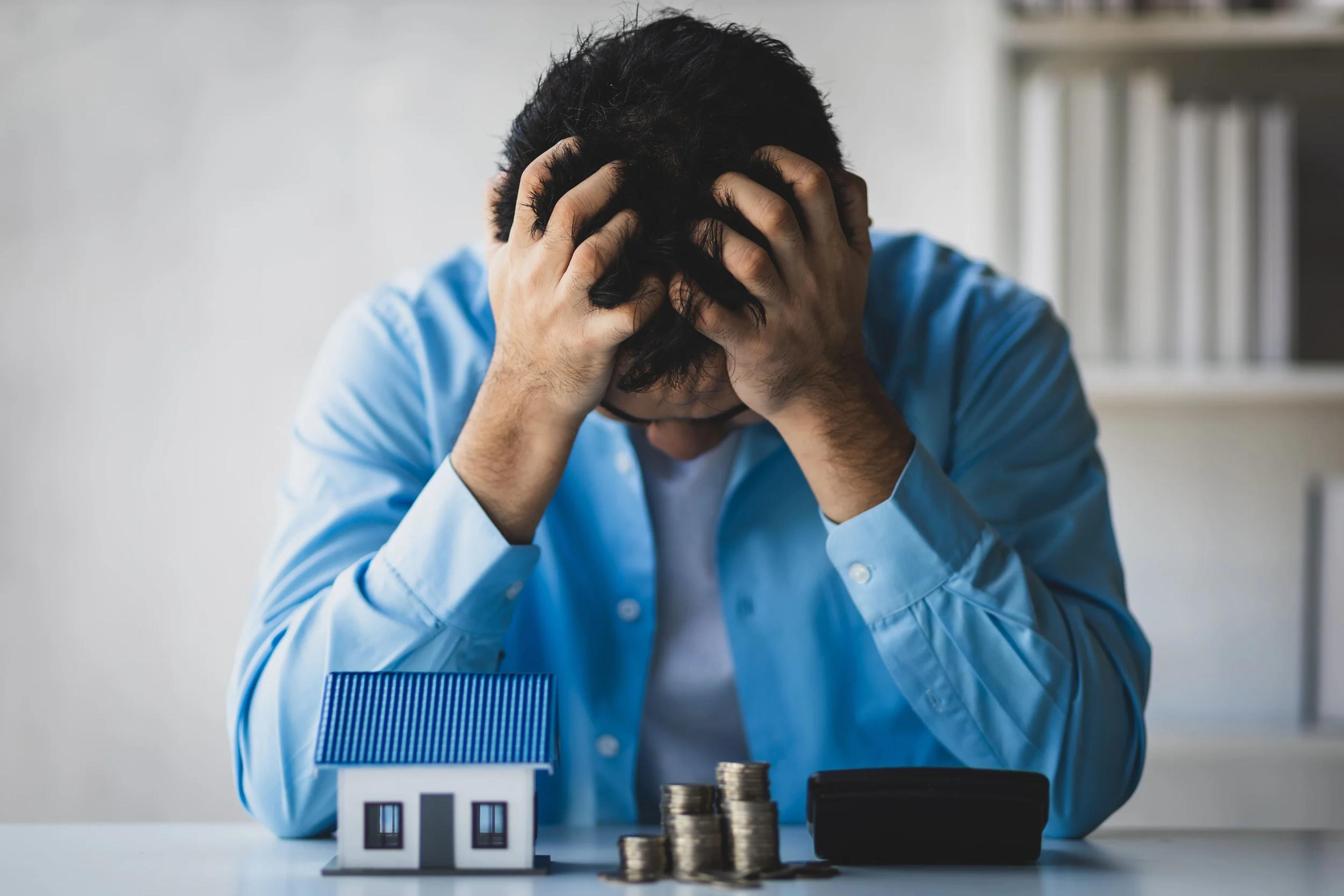 Stressed man with his head in his hands beside a house model and coins after an investment mistake