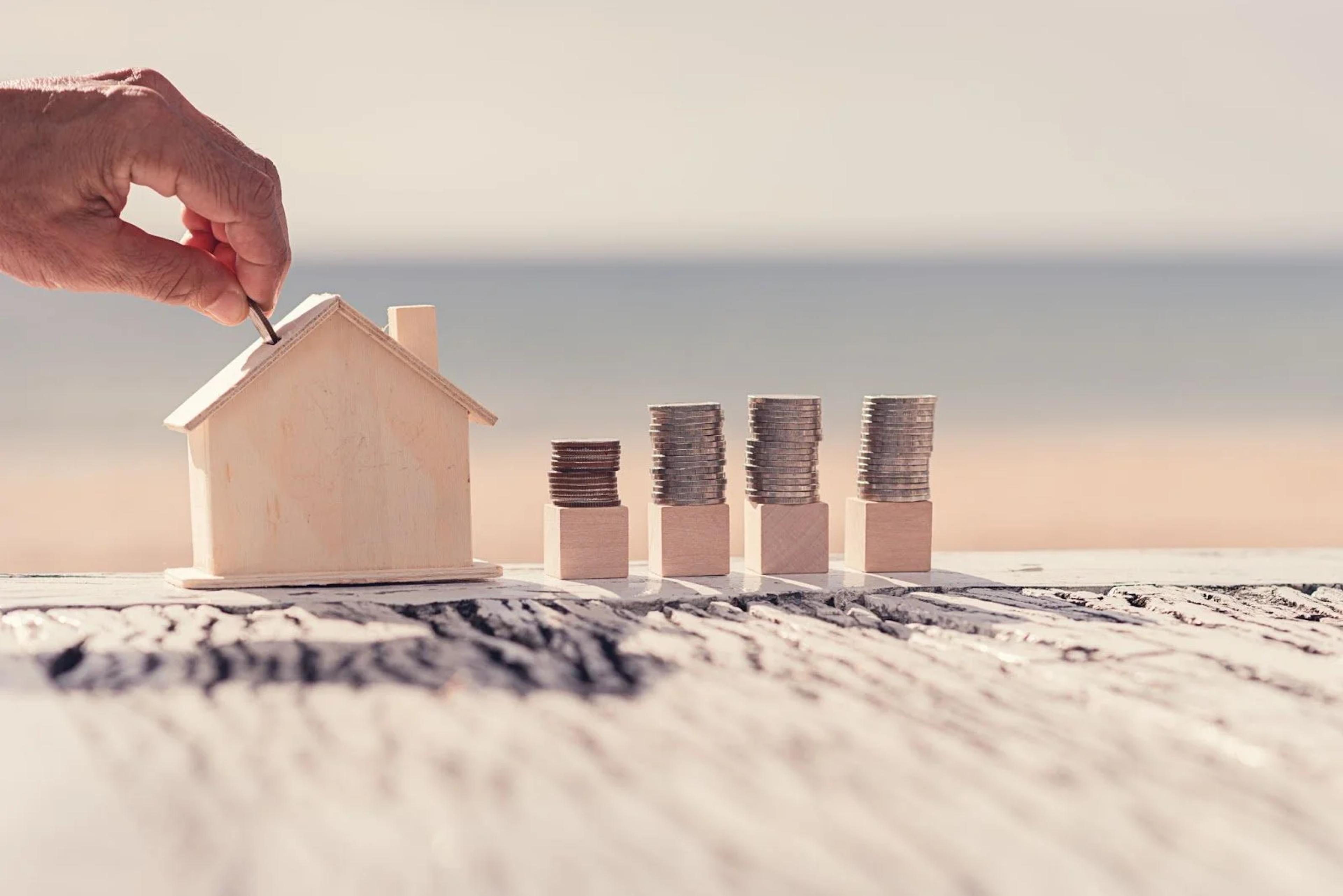 Hand dropping a coin into a wooden house-shaped money box beside coin stacks