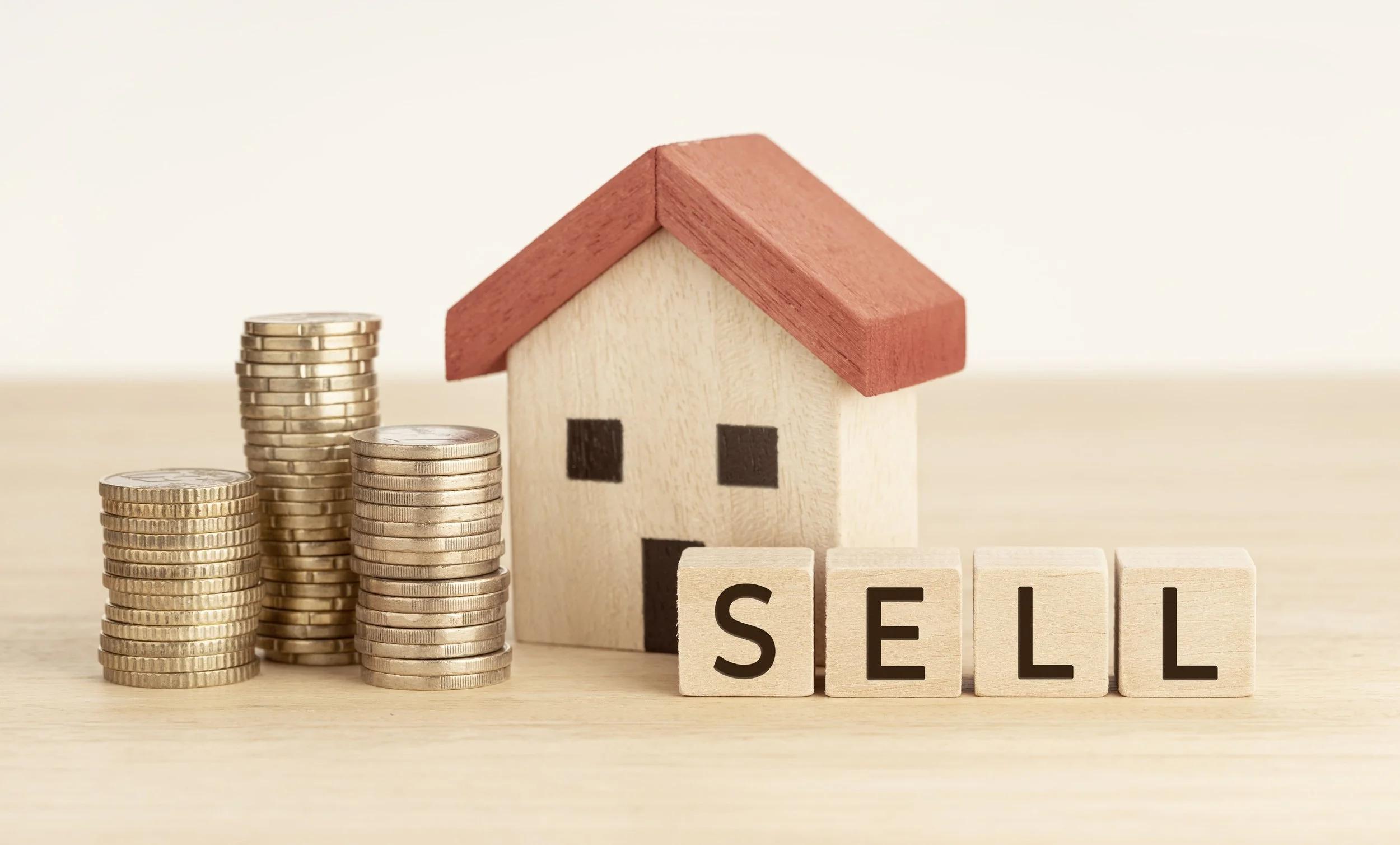 House model beside stacks of coins and wooden blocks spelling SELL