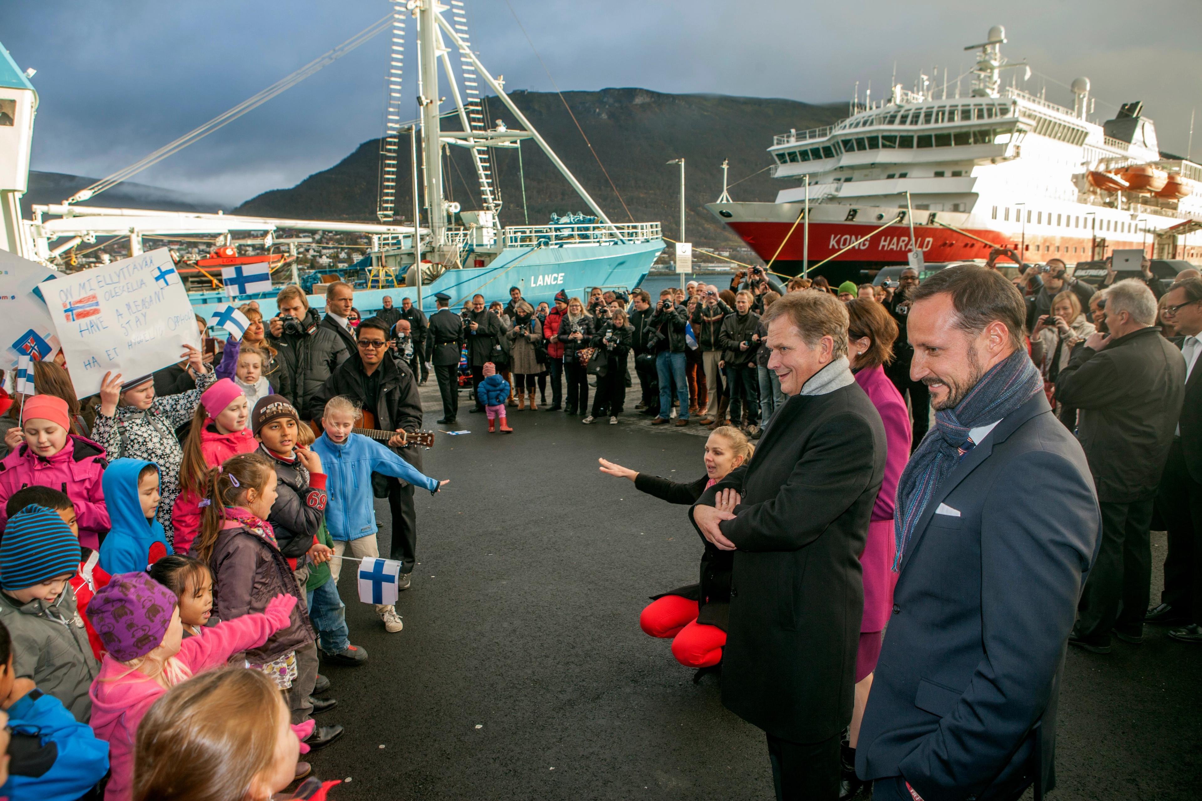 Presidenten og Kronprinsen står smilende foran en flokk små barn. To skip ligger ved kai i bakgrunnen.
