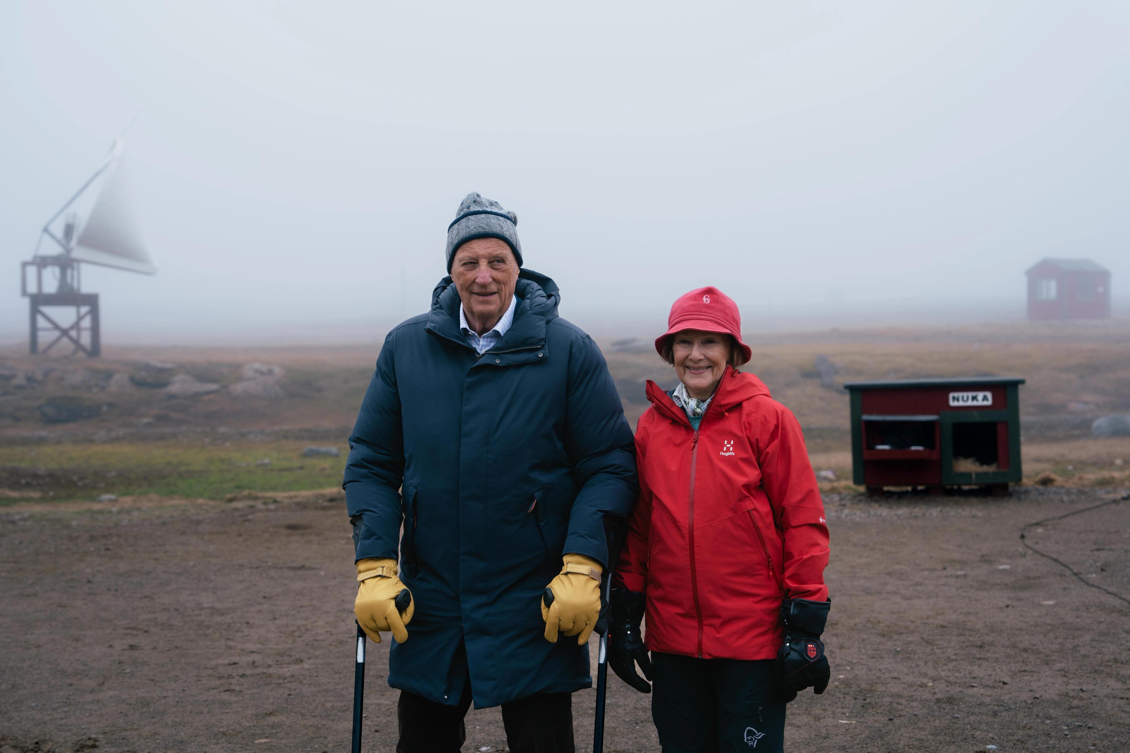 Kong Harald og Dronning Sonja står godt kledd ute. Det er tåke, og i bakgrunnen skimtes et hundehus og en parabol.