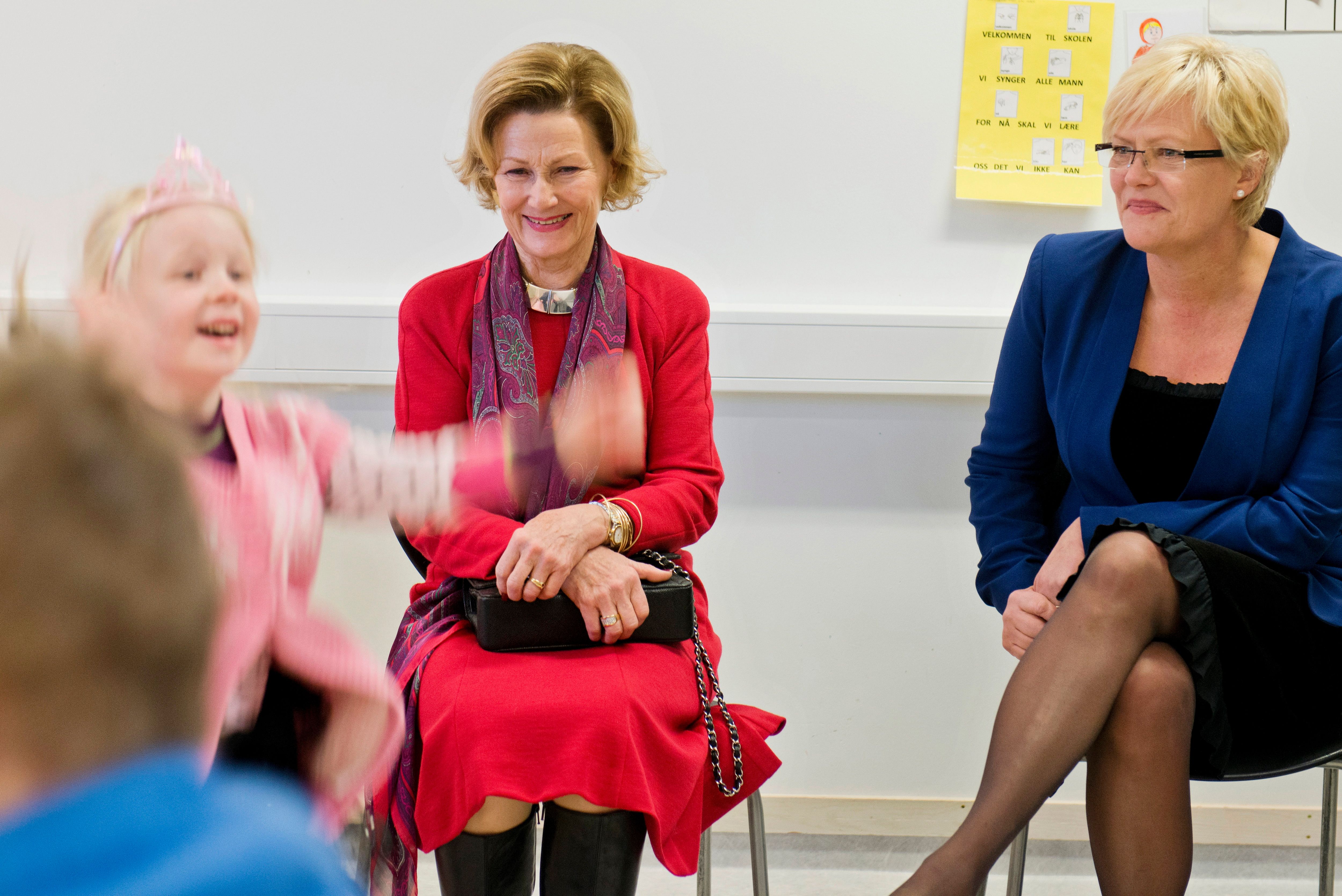 Dronning Sonja sitter på en stol i et klasserom under besøket til Fagerlund skole. Hun smiler, mens vi ser en ung jente i rosa klær og leketiara vifte med hendene.