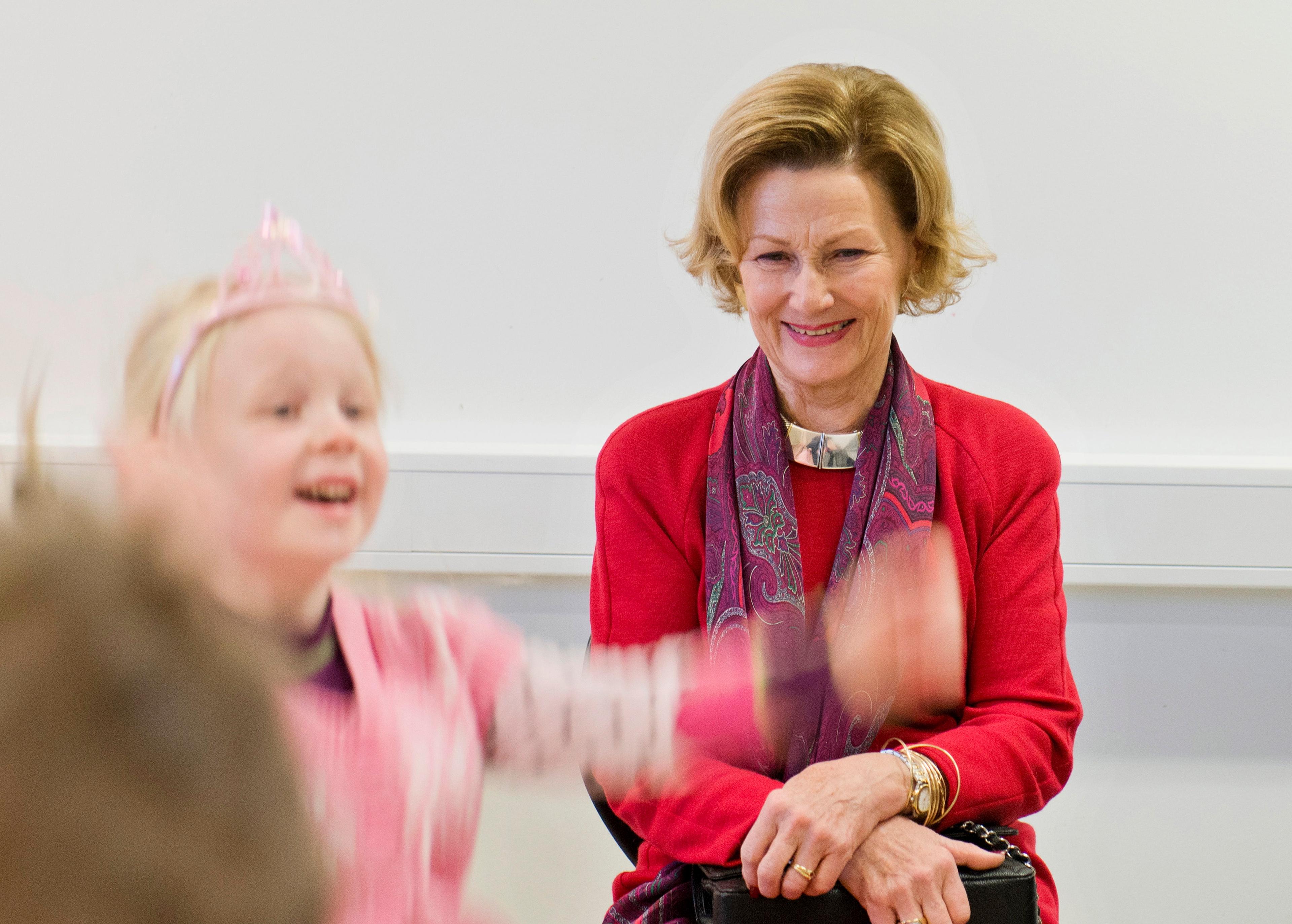 Dronning Sonja sitter på en stol i et klasserom under besøket til Fagerlund skole. Hun smiler, mens vi ser en ung jente i rosa klær og leketiara vifte med hendene.