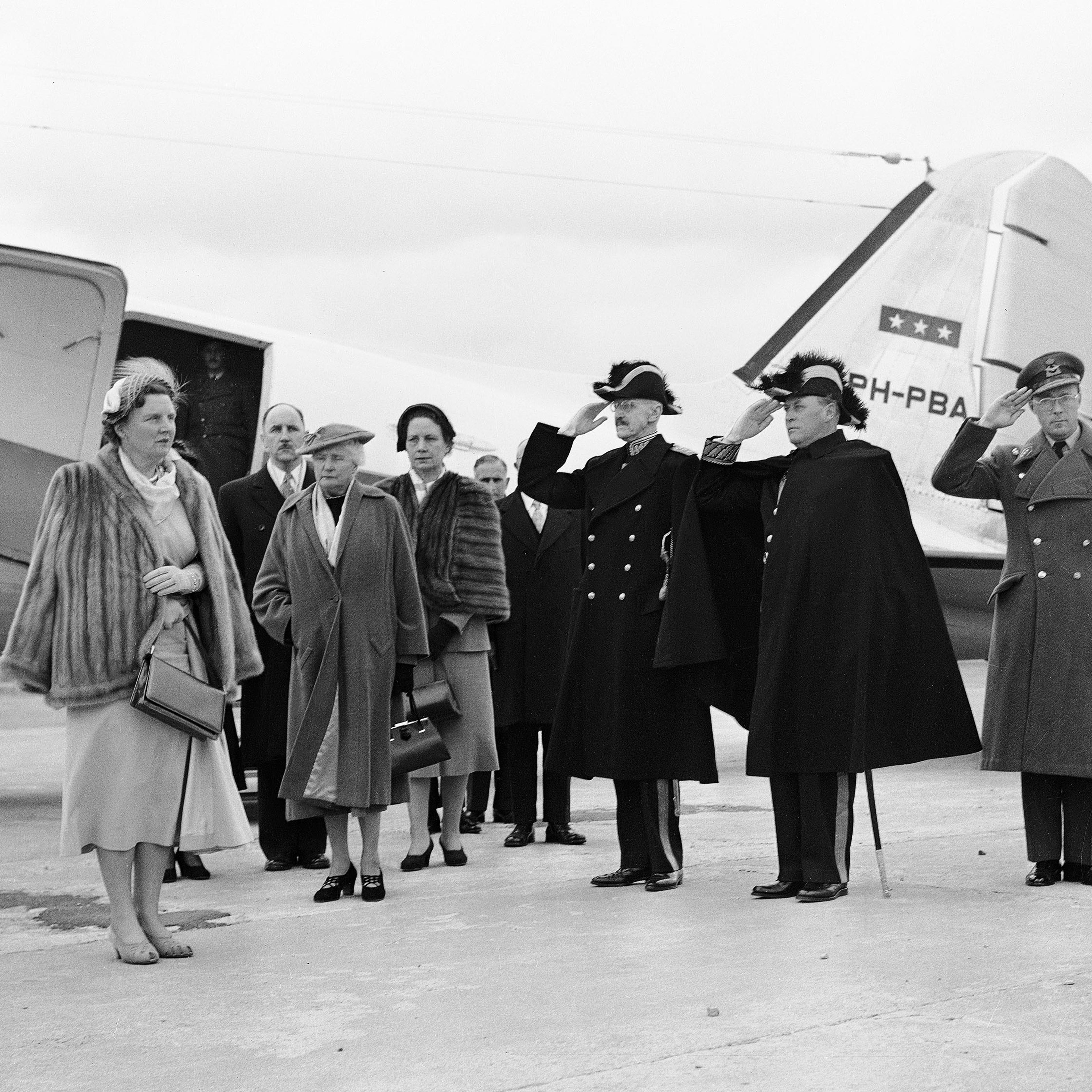 Dronning Juliana ønskes velkommen utenfor flyet. Kong Haakon og Kronprins Olav gjør honnør. Dronning Juliana har pelsjakke og hatt. Foto: Valldal NTB
