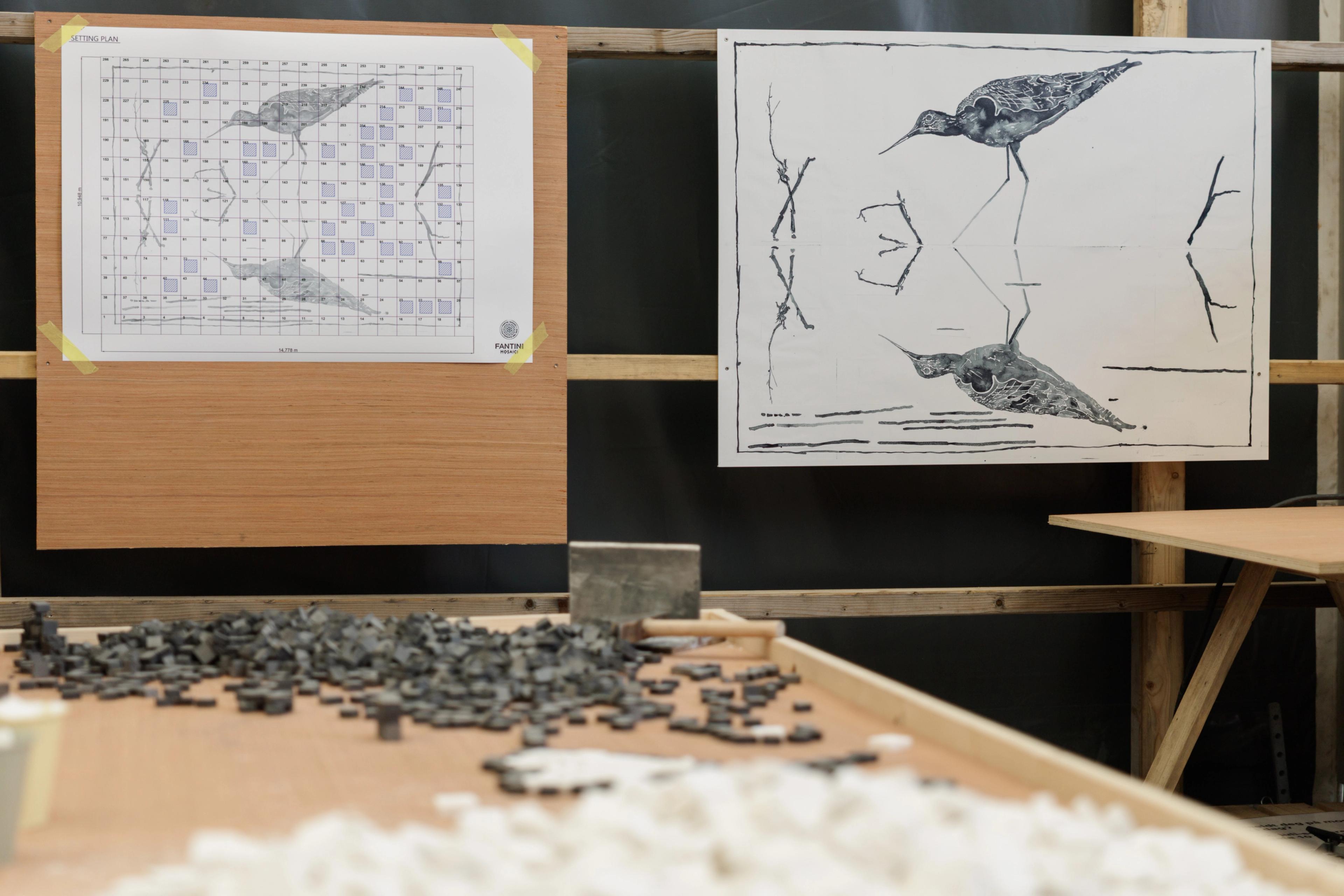 I bakgrunnen er en tegning av det ferdige kunstverket og arbeidstegning hengt opp. På et bord i forgrunnen ligger sorte og hvite mosaikksteiner klare til å tas i bruk.