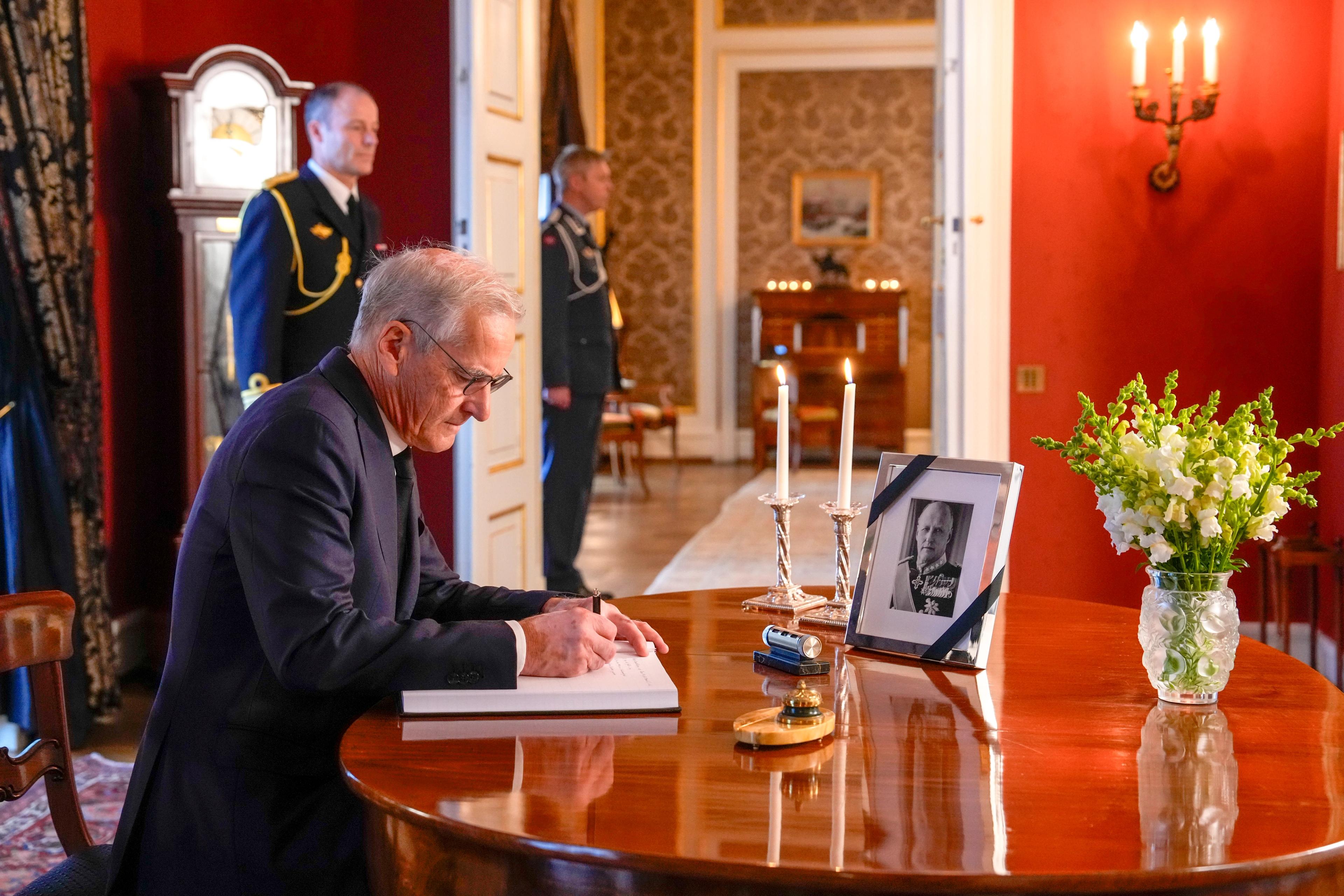 Støre sitter ved et bord og skriver. På bordet står et bilde av Kong Harald med sørgeflor omkring rammen og en bukett hvite blomster.