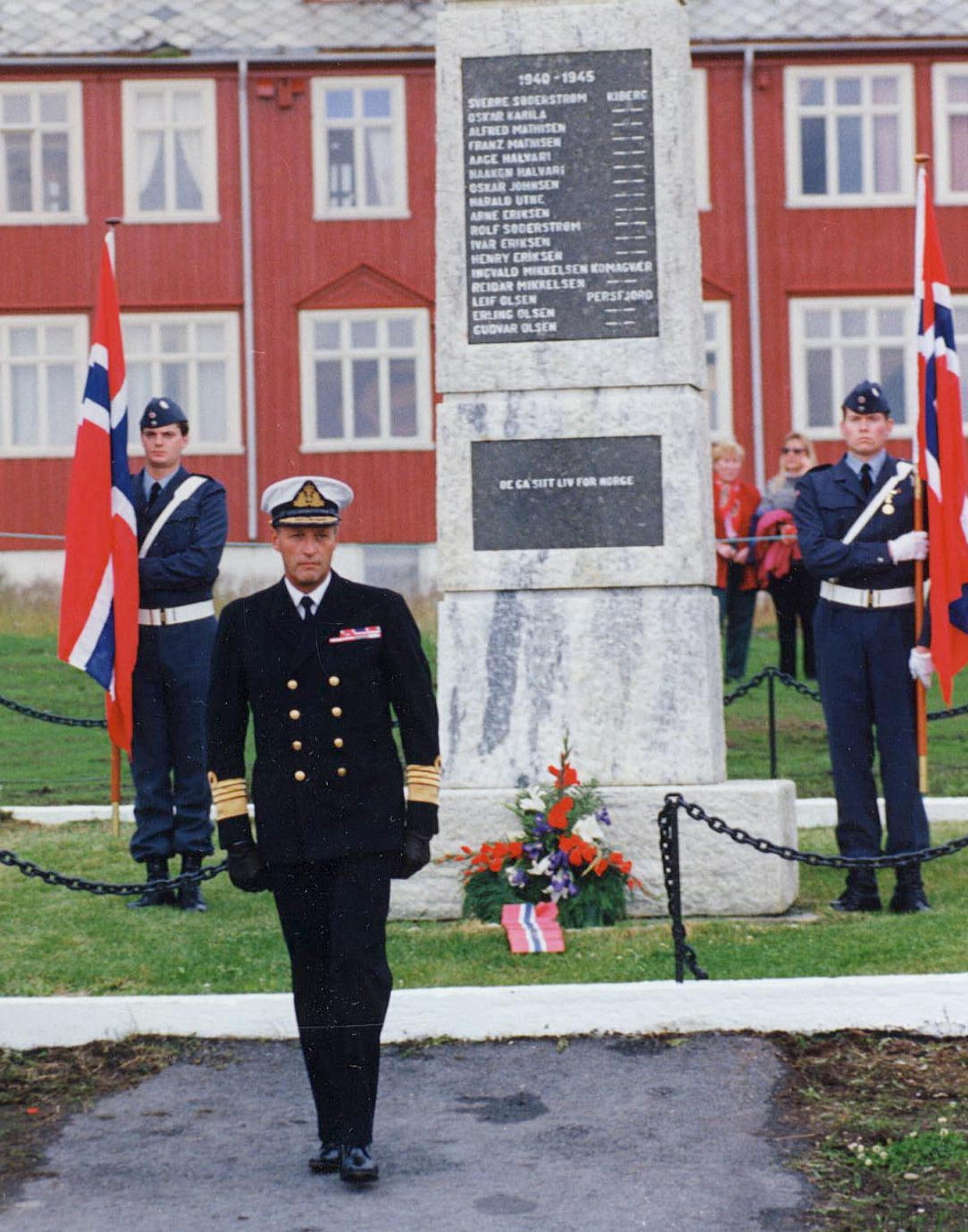 Kongen i marineuniform foran bautaen med de falnes navn. Soldater med store norske flagg på hver side av bautaen.