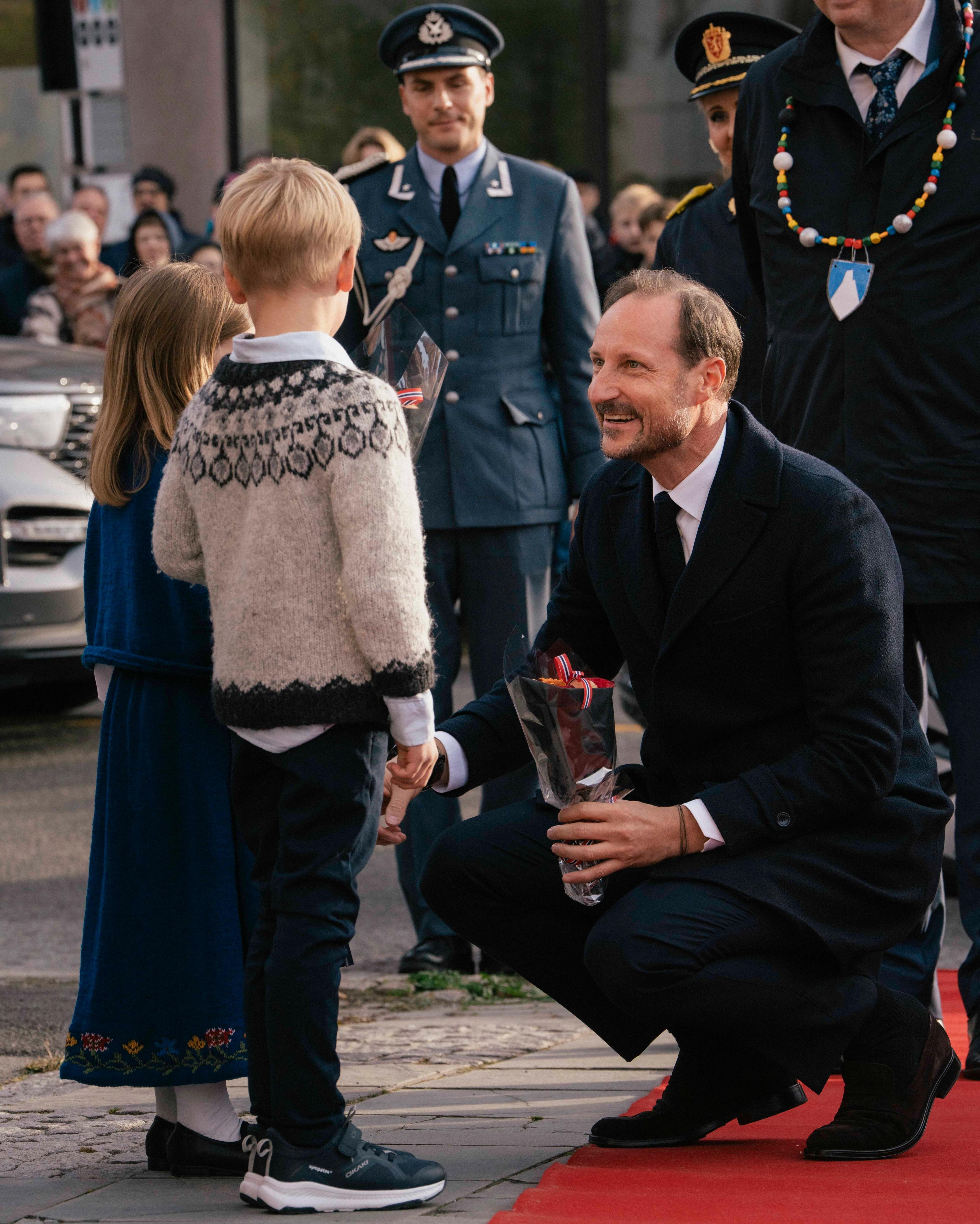 Kong Haakon sitter på huk på den røde løperen foran to små barn.