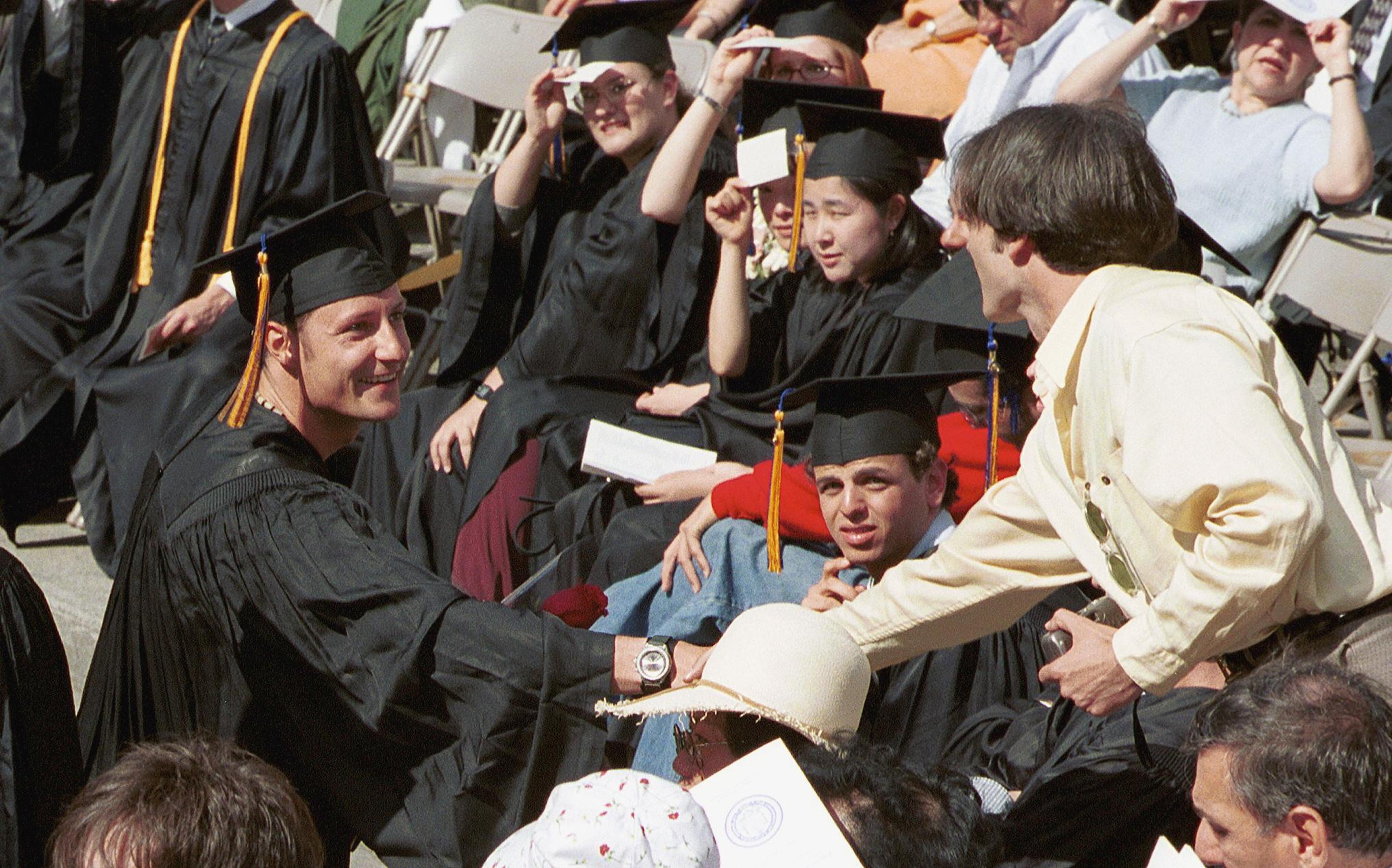 Kronprins Haakon er ikledd bachelorkappe og hatt og hilser på en person i sivil.