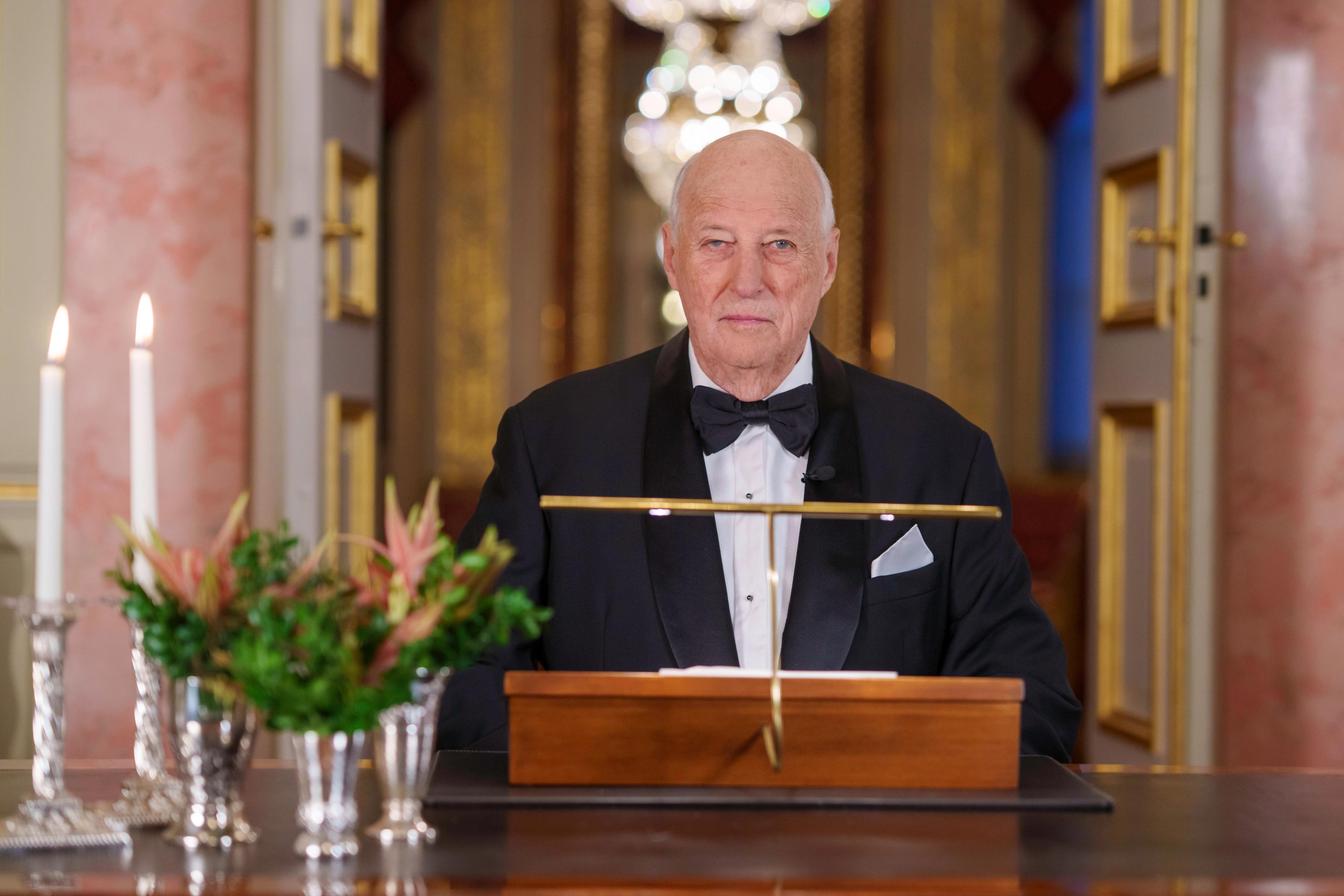 Kong Harald sitter ved skrivebordet med talen foran seg. Han er kledd i smoking. Det er levende lys og blomster på bordet foran ham.