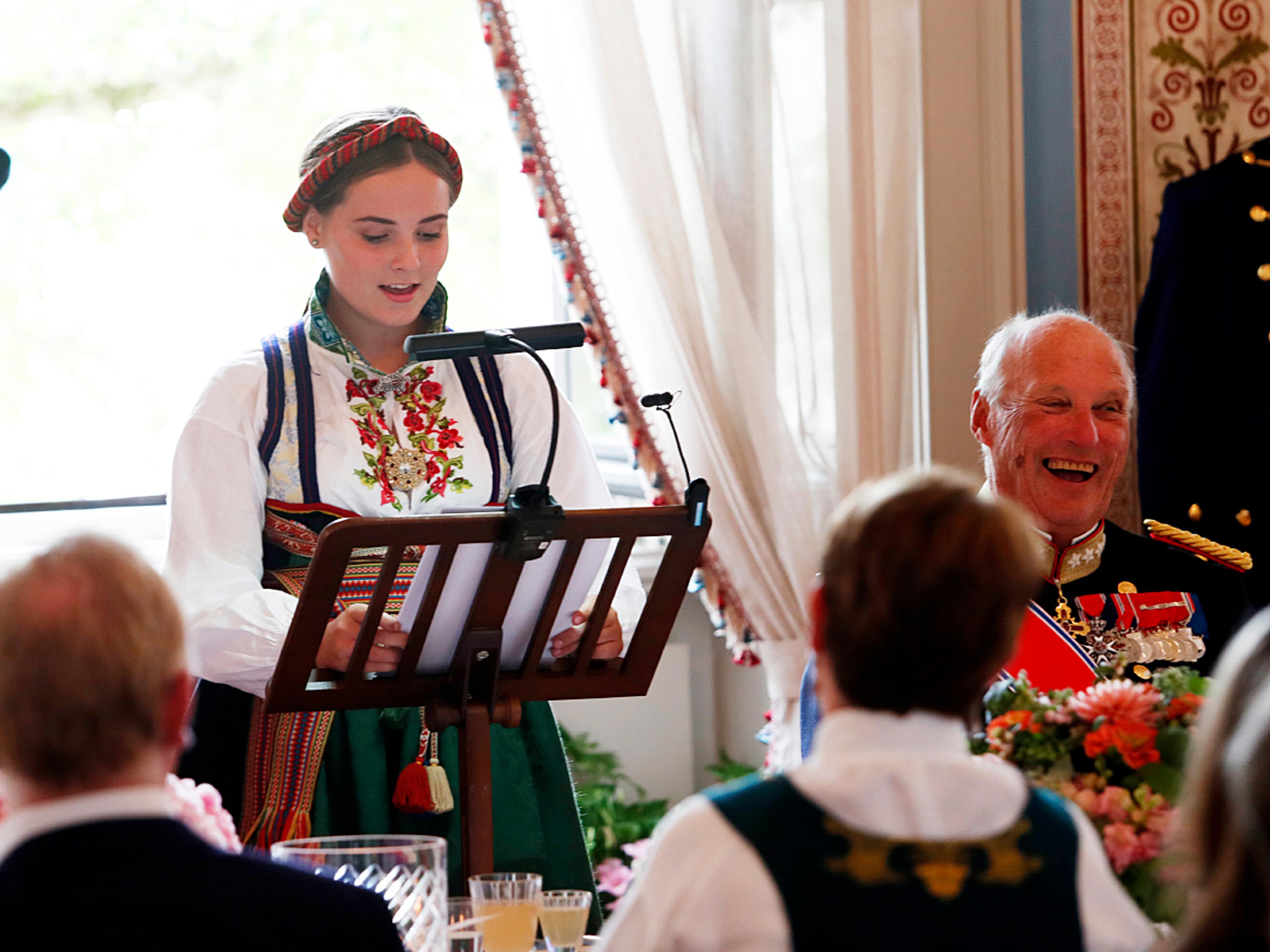 Prinsesse Ingrid Alexandra står i bunad ved bordet og taler. Kongen sitter ved siden av og ler godt av noe hun sier.